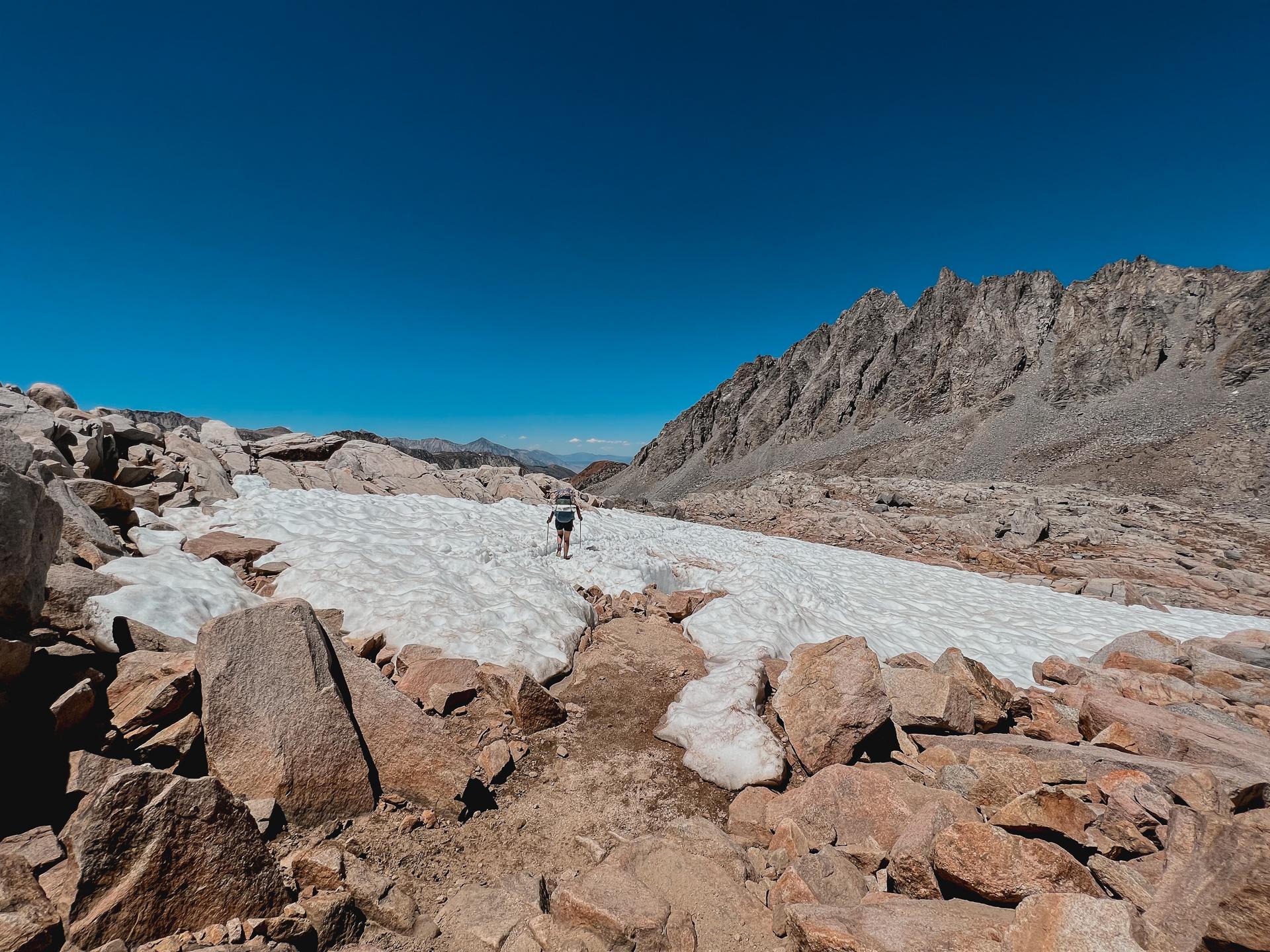Hiker walks across a snow patch in a rocky mountain landscape.