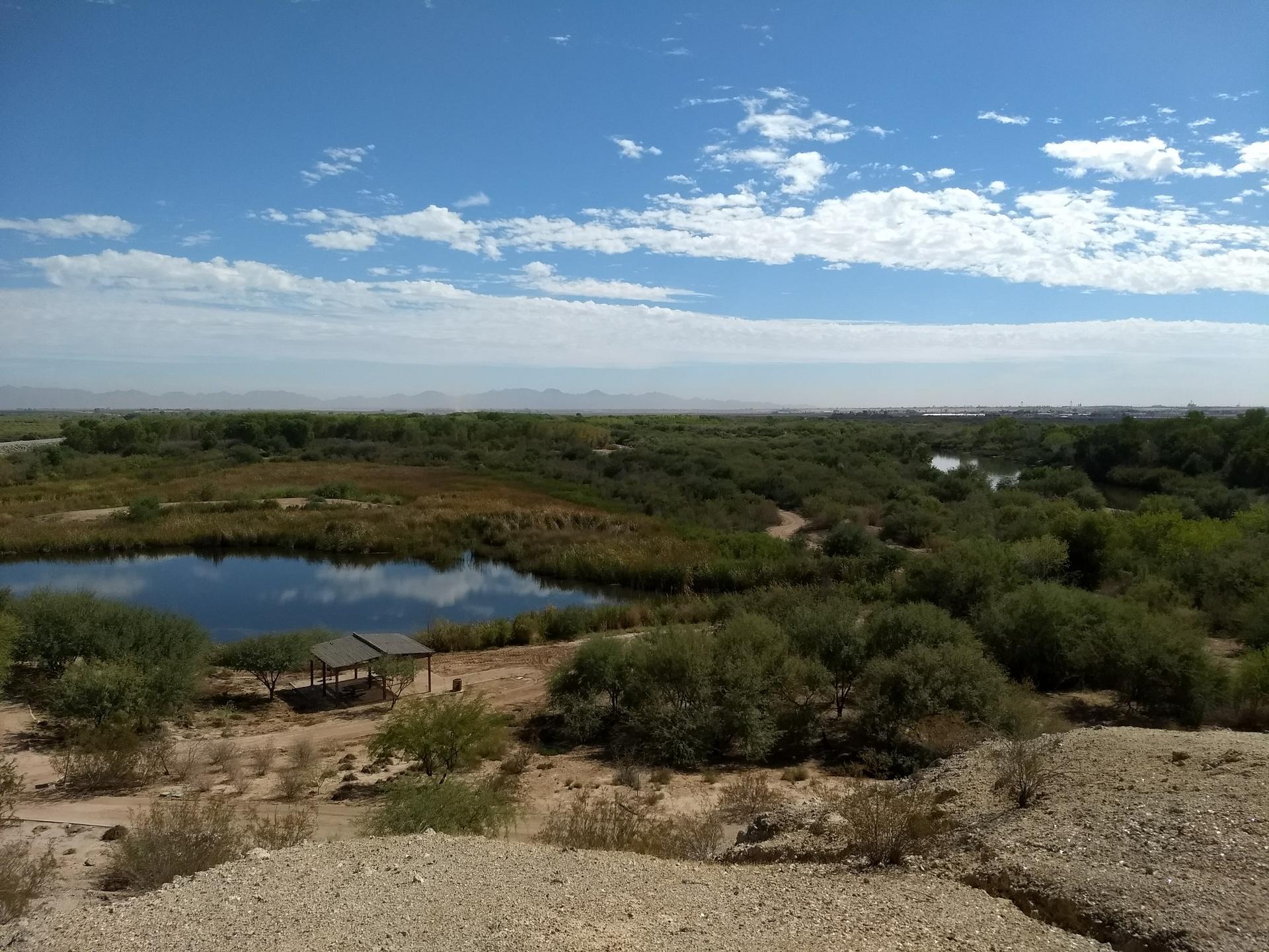 Expansive green landscape with a pond, river, and shelter under a blue sky.