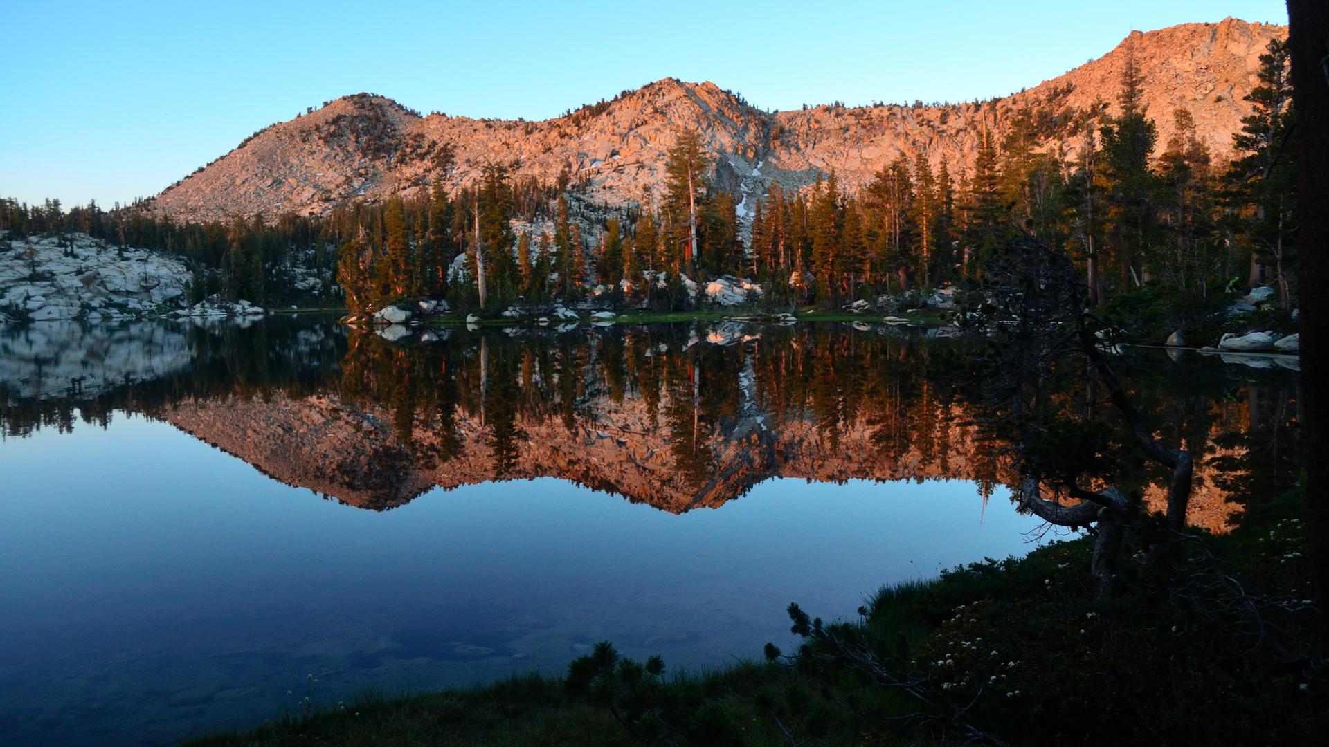 Warm light on mountains and trees reflected in a still lake at dawn.