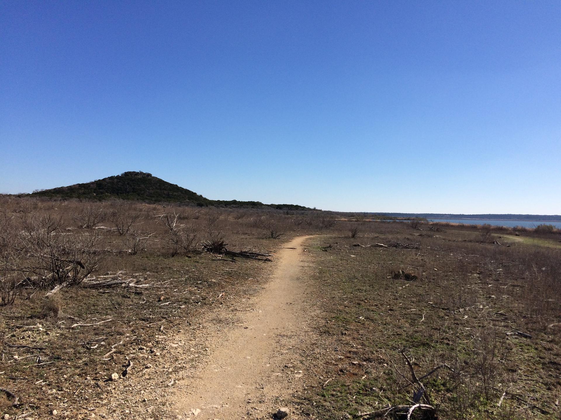 Dirt trail through dry, sparse landscape with a distant hill and water, clear sky.