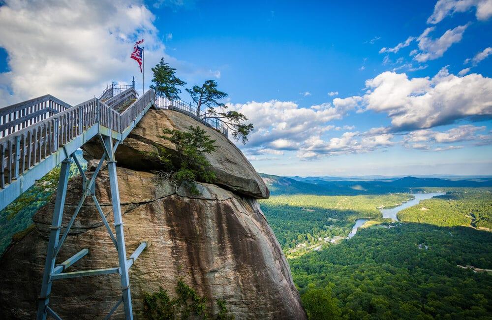 Metal staircase leading to a rocky peak with an American flag, overlooking a river. Bright daylight.