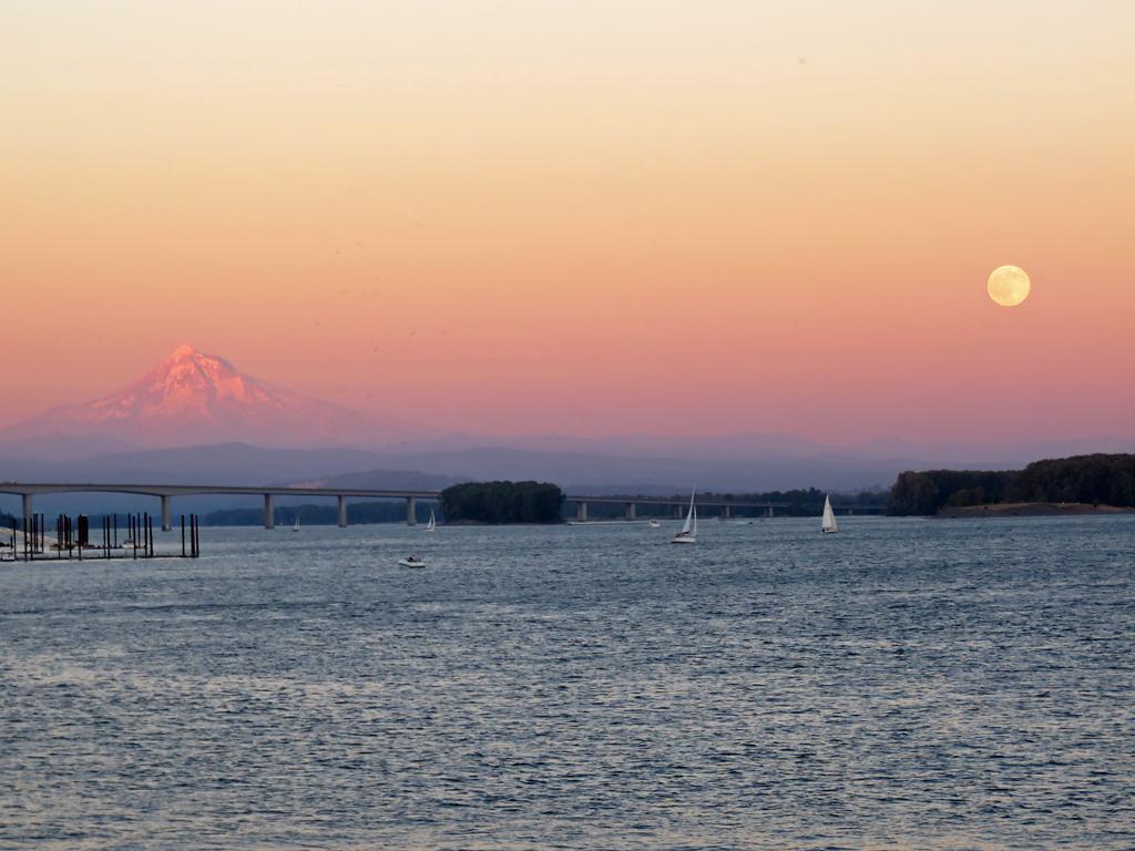 Sunset over a river with a full moon, distant mountain, and sailboats.