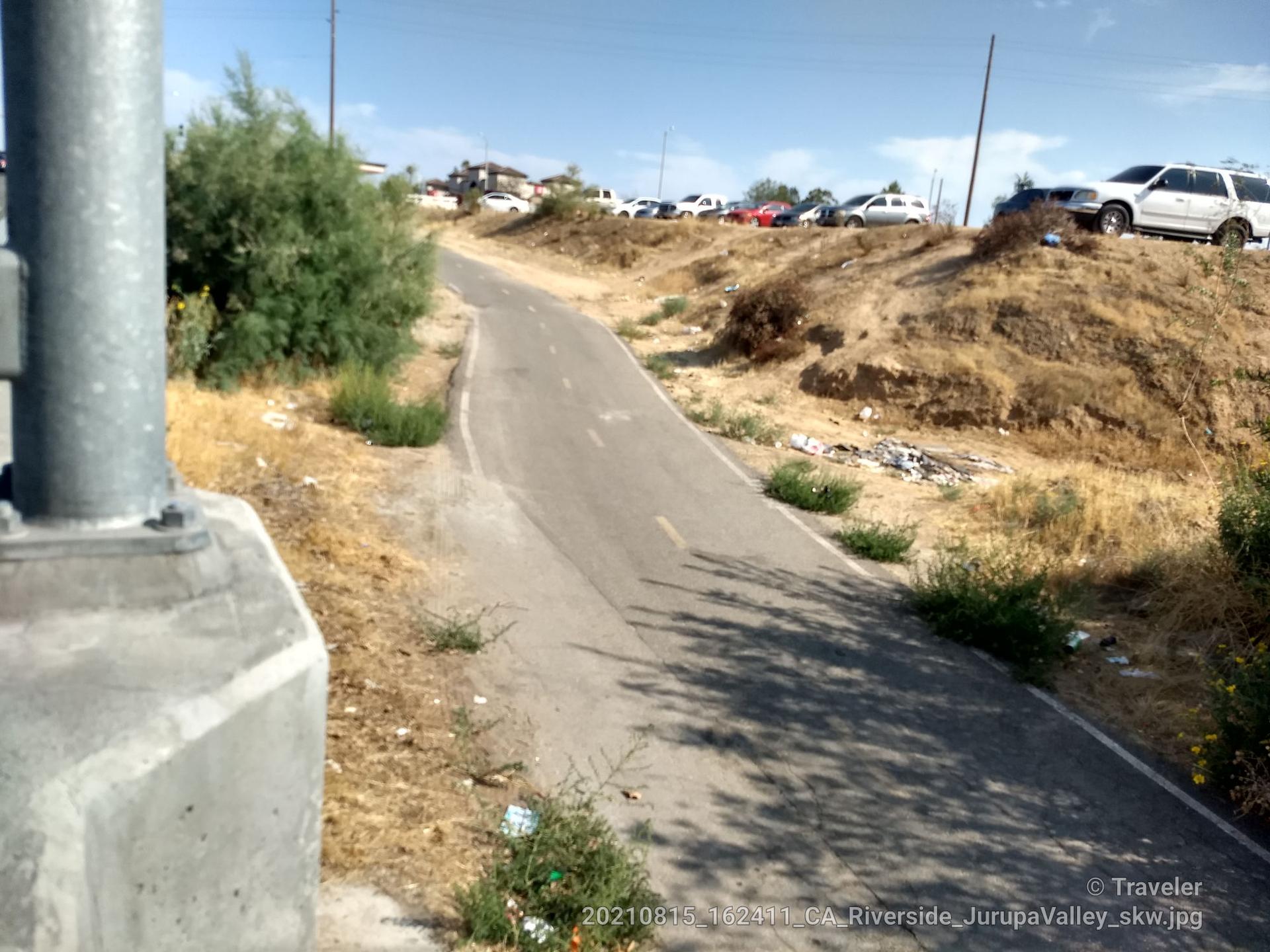 Paved path winding uphill beside dry vegetation, with parked cars in the distance.
