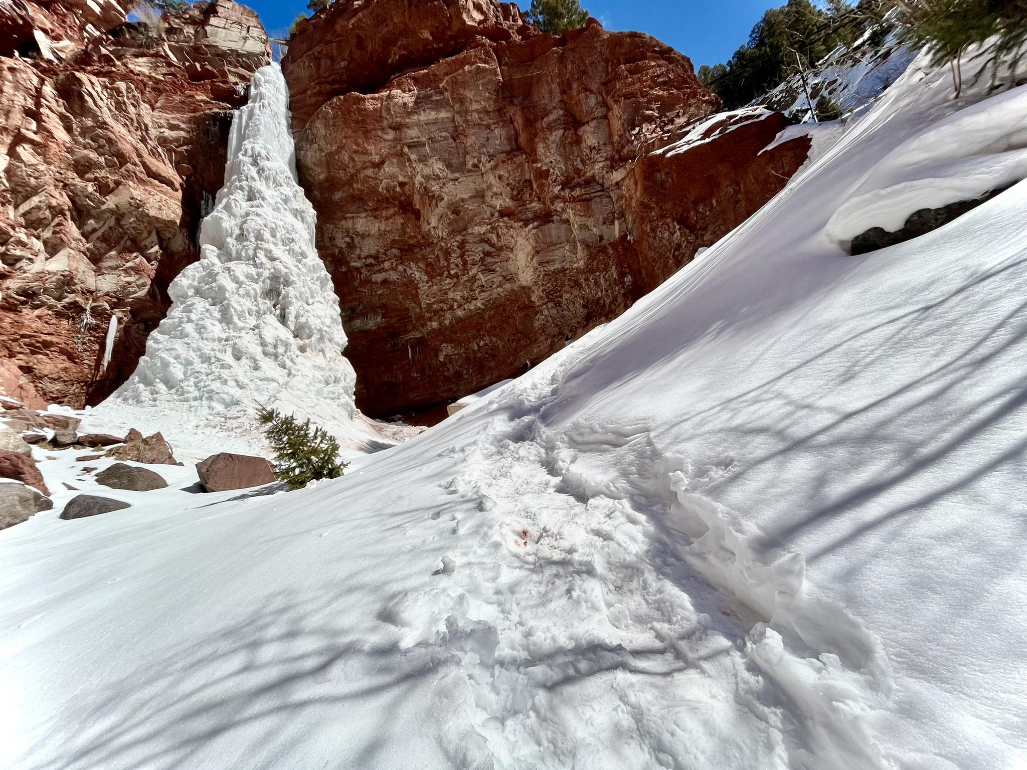 A massive frozen waterfall stands between red cliffs, with snowy footprints on a sunny slope.