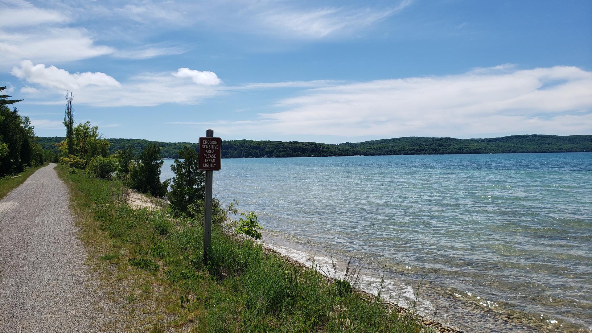 A gravel path runs beside a large blue lake under a sunny sky.