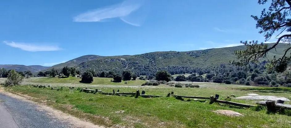 Wide green field with a wooden fence, backed by mountains under a blue sky.