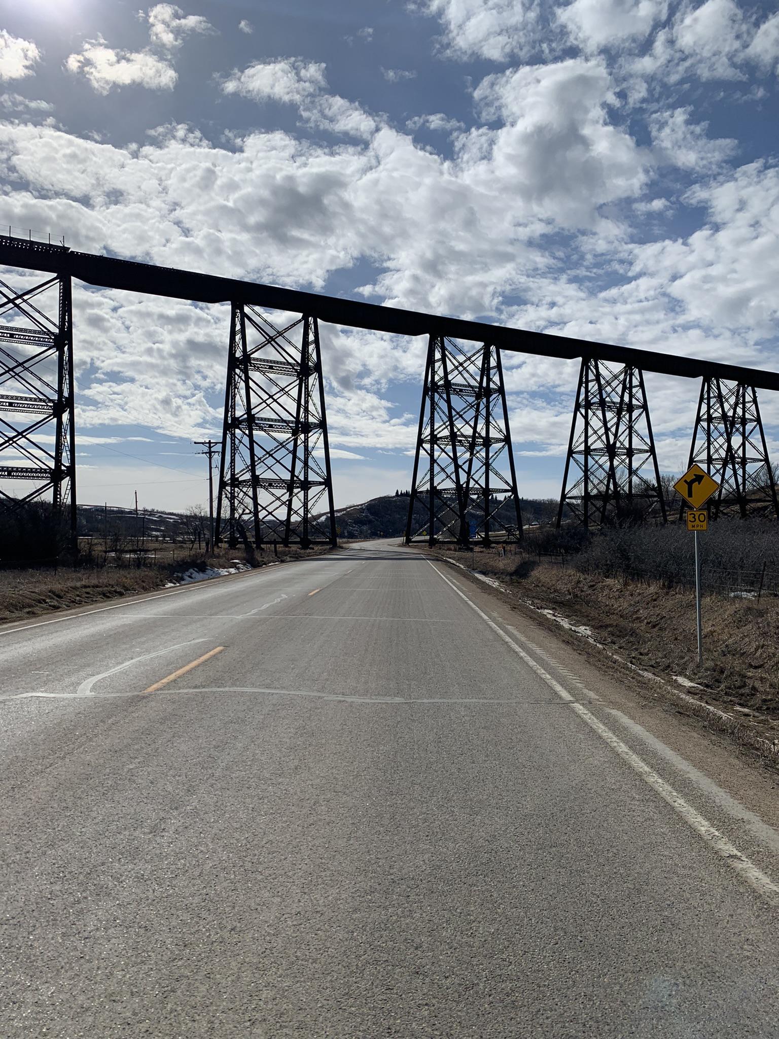 A long, dark metal trestle bridge crosses above a sunlit road.