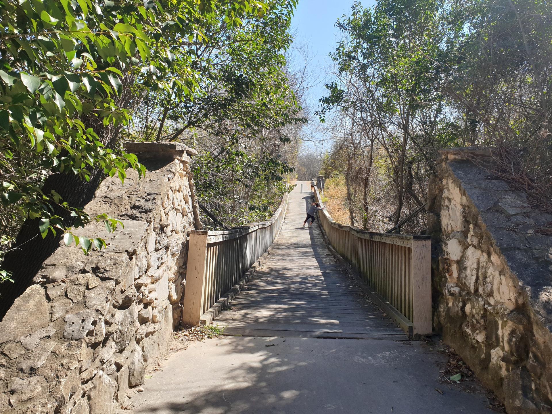 Stone and wood bridge over a path with lush green trees, person walking.