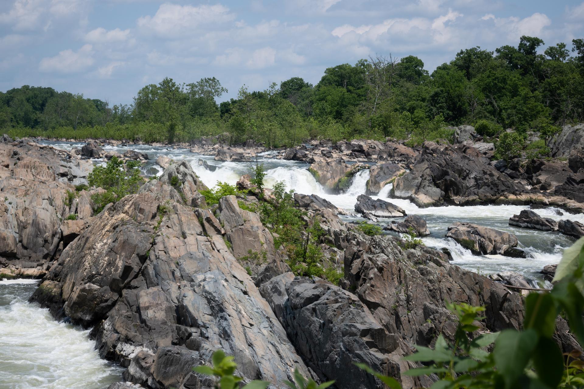 River rapids flowing through rocky formations, surrounded by green trees under cloudy sky.
