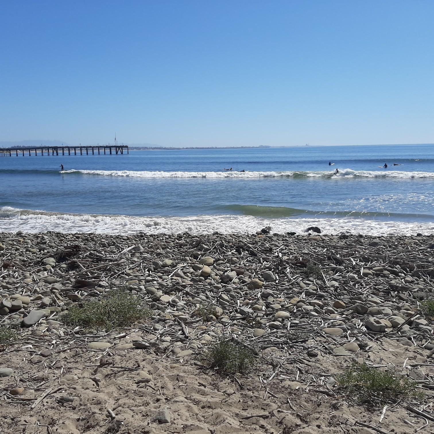 Rocky beach with ocean waves, distant surfers, and a long pier under blue sky.