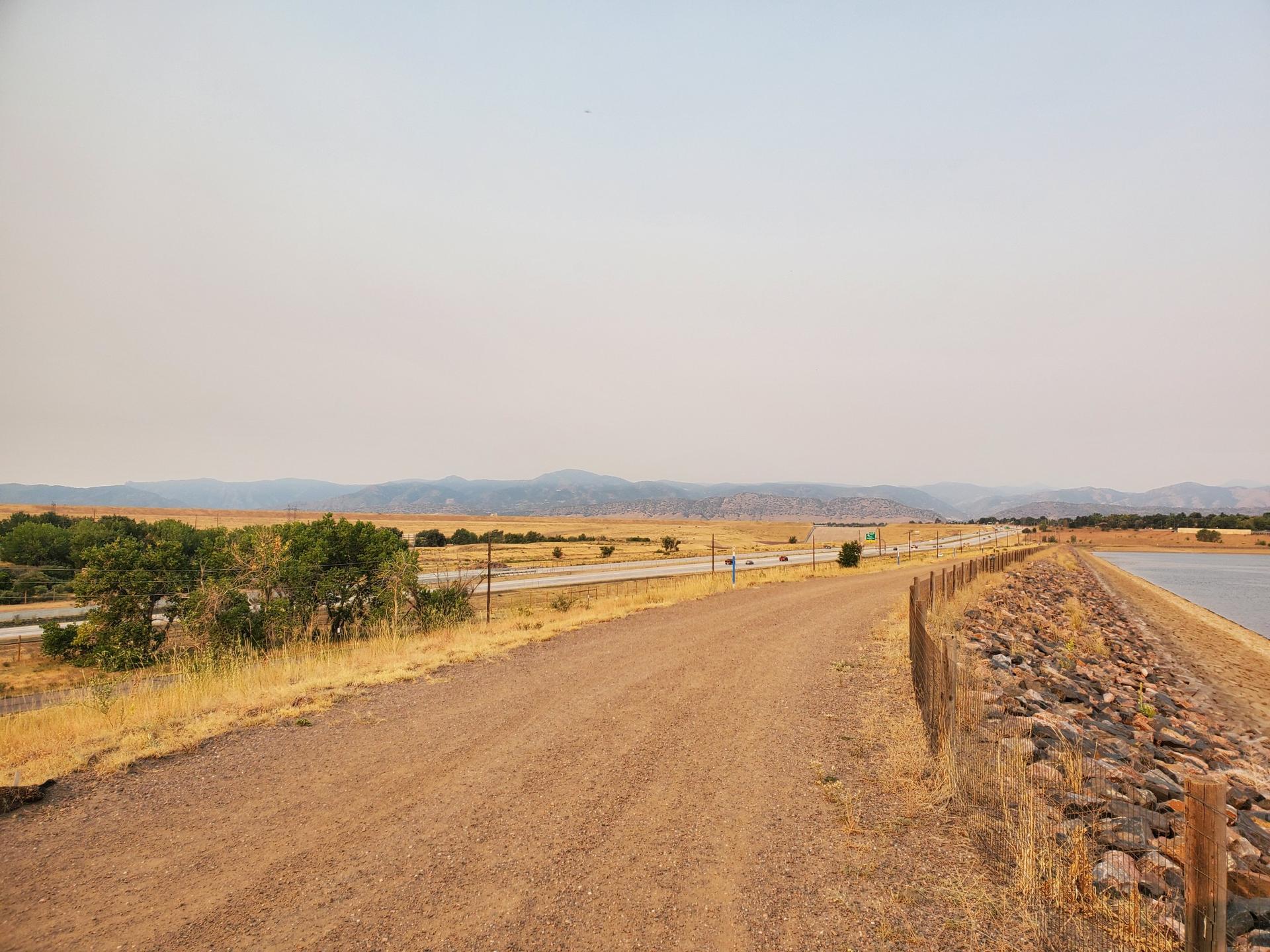 A dirt road next to a highway and reservoir, with hazy mountains in distance.
