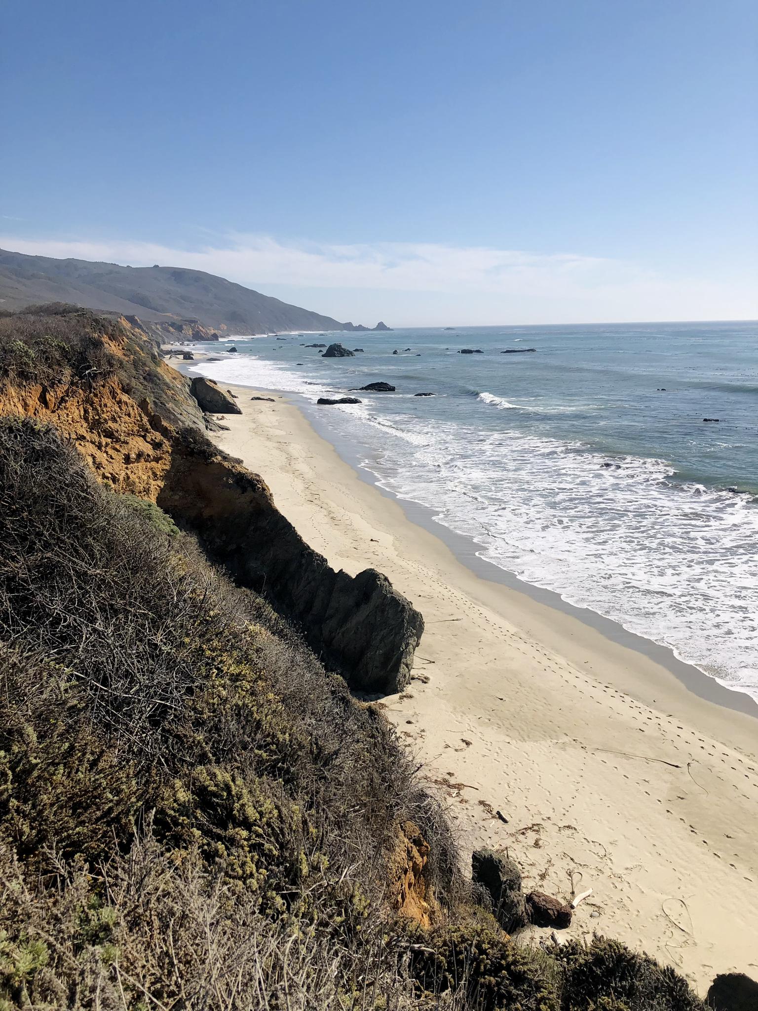 Bright sunny view of a sandy beach with waves and a steep coastal cliff.