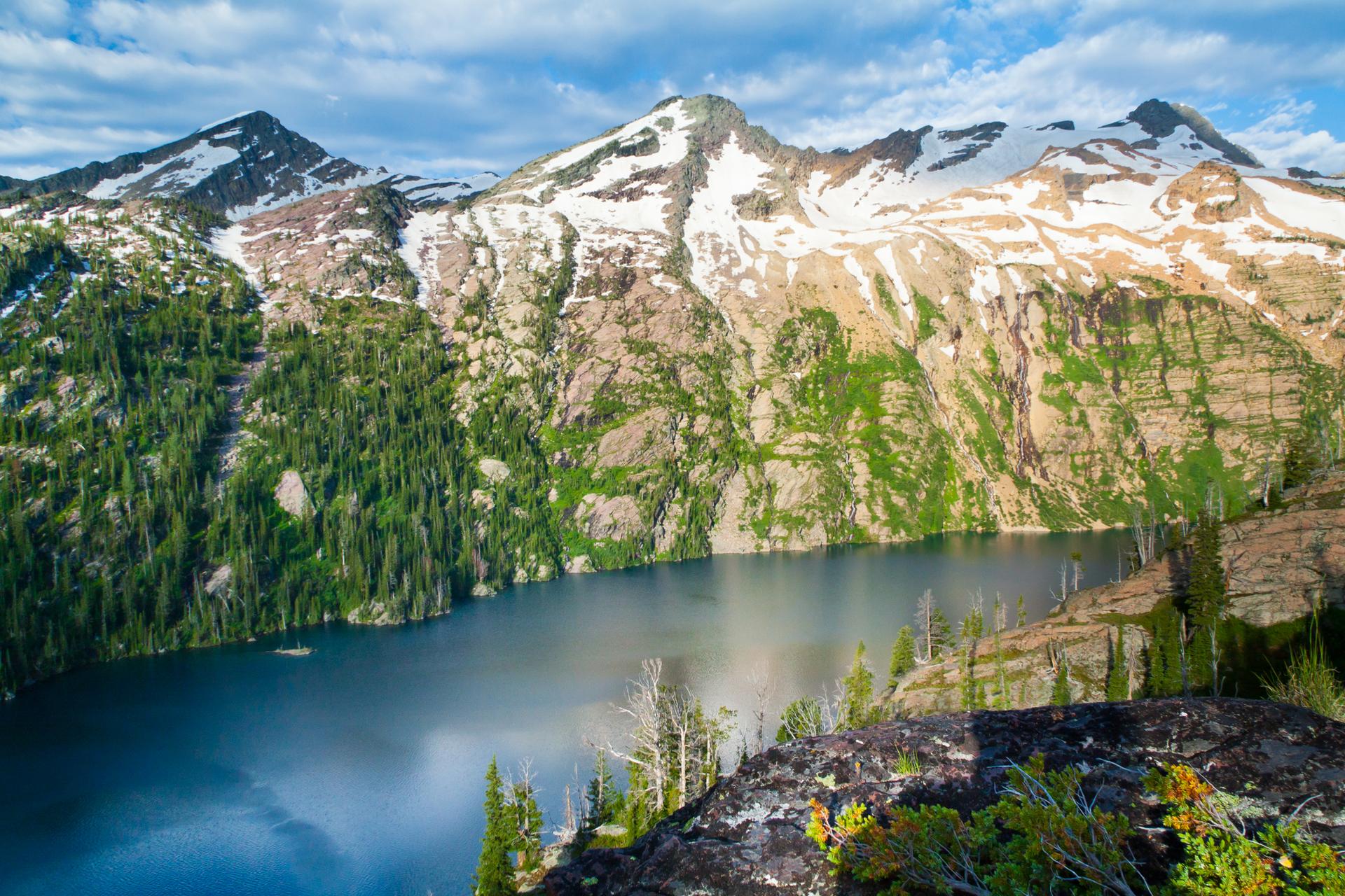 Vast alpine lake surrounded by snow-capped mountains and dense green forests under blue sky.