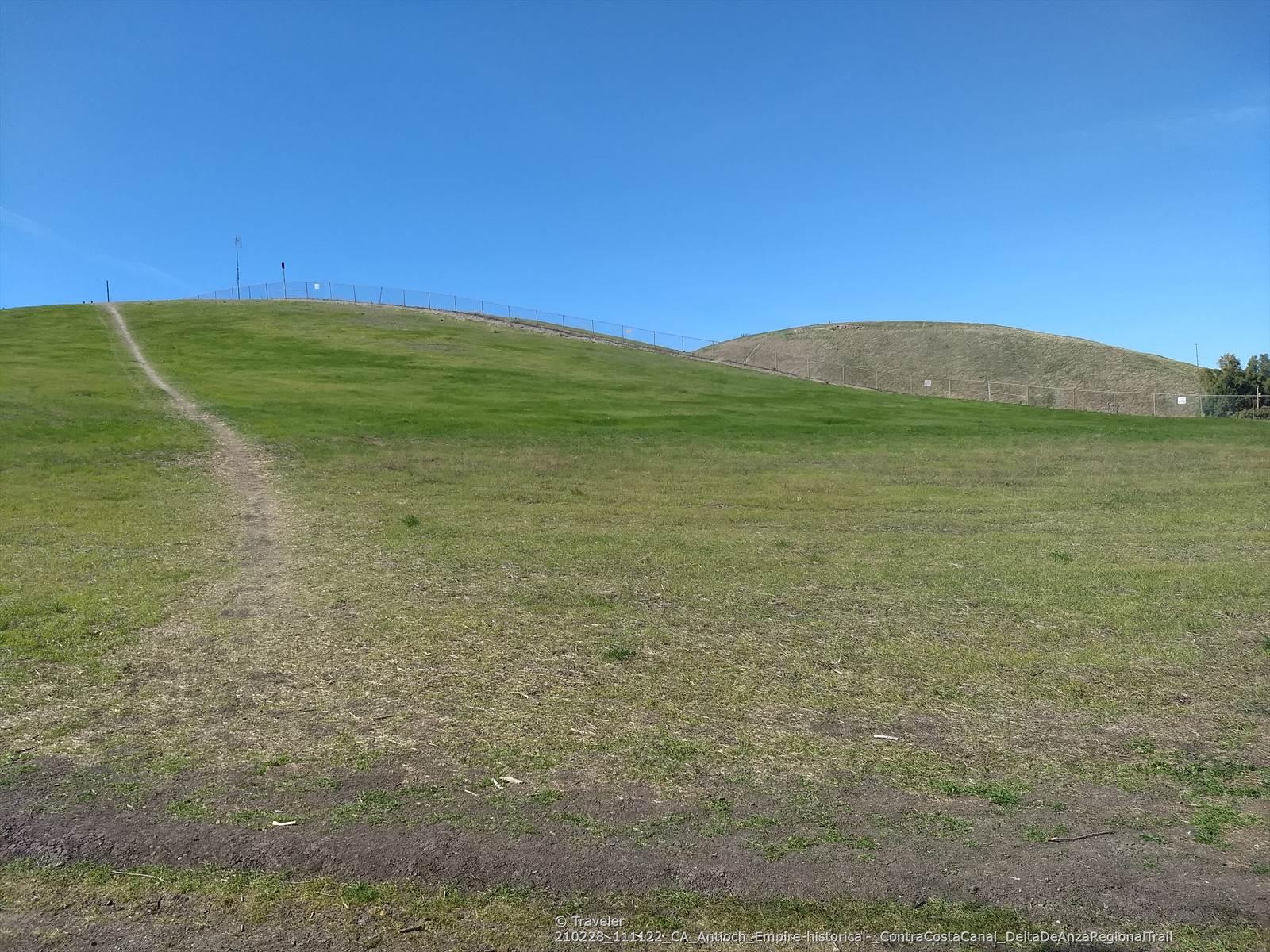 Green grassy hills with a dirt path leading upwards under a clear blue sky.