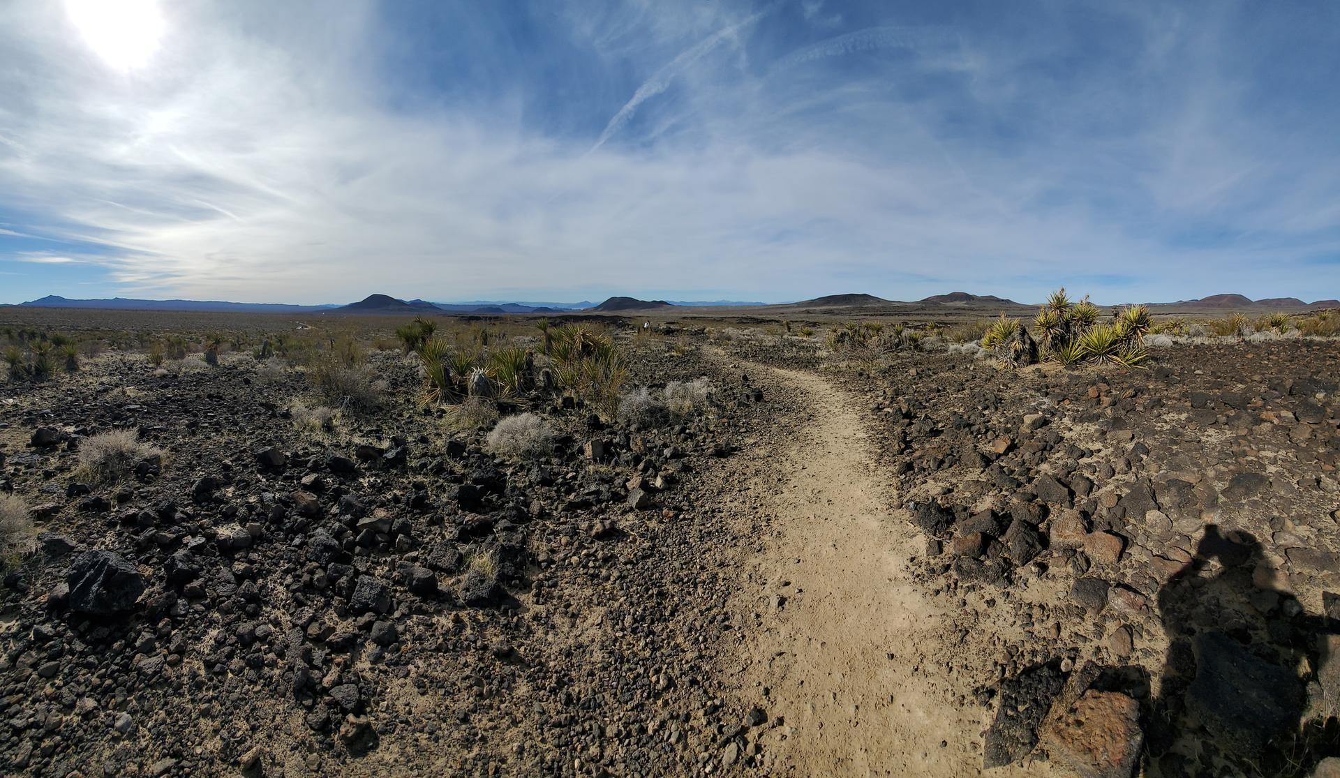 A dirt path winds through a vast, rocky desert under a bright sky.