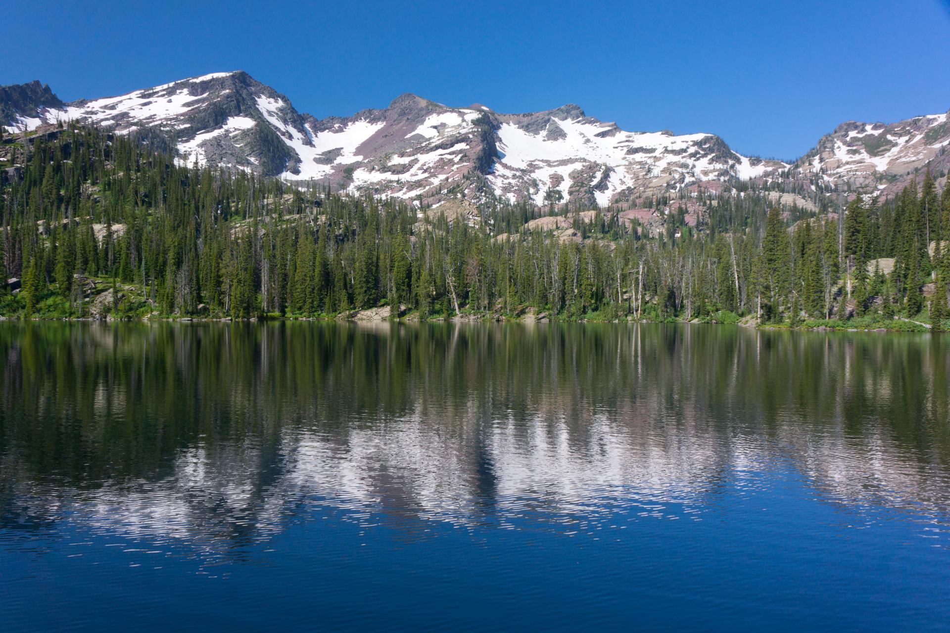 Clear lake reflects snow-capped mountains and dense evergreen forest on a bright day.