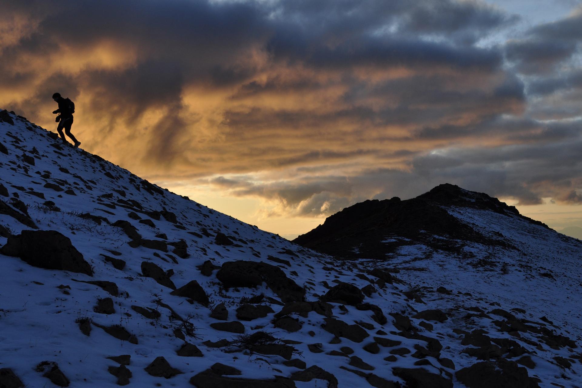 Silhouetted person hiking up a snowy, rocky mountain slope at sunset.