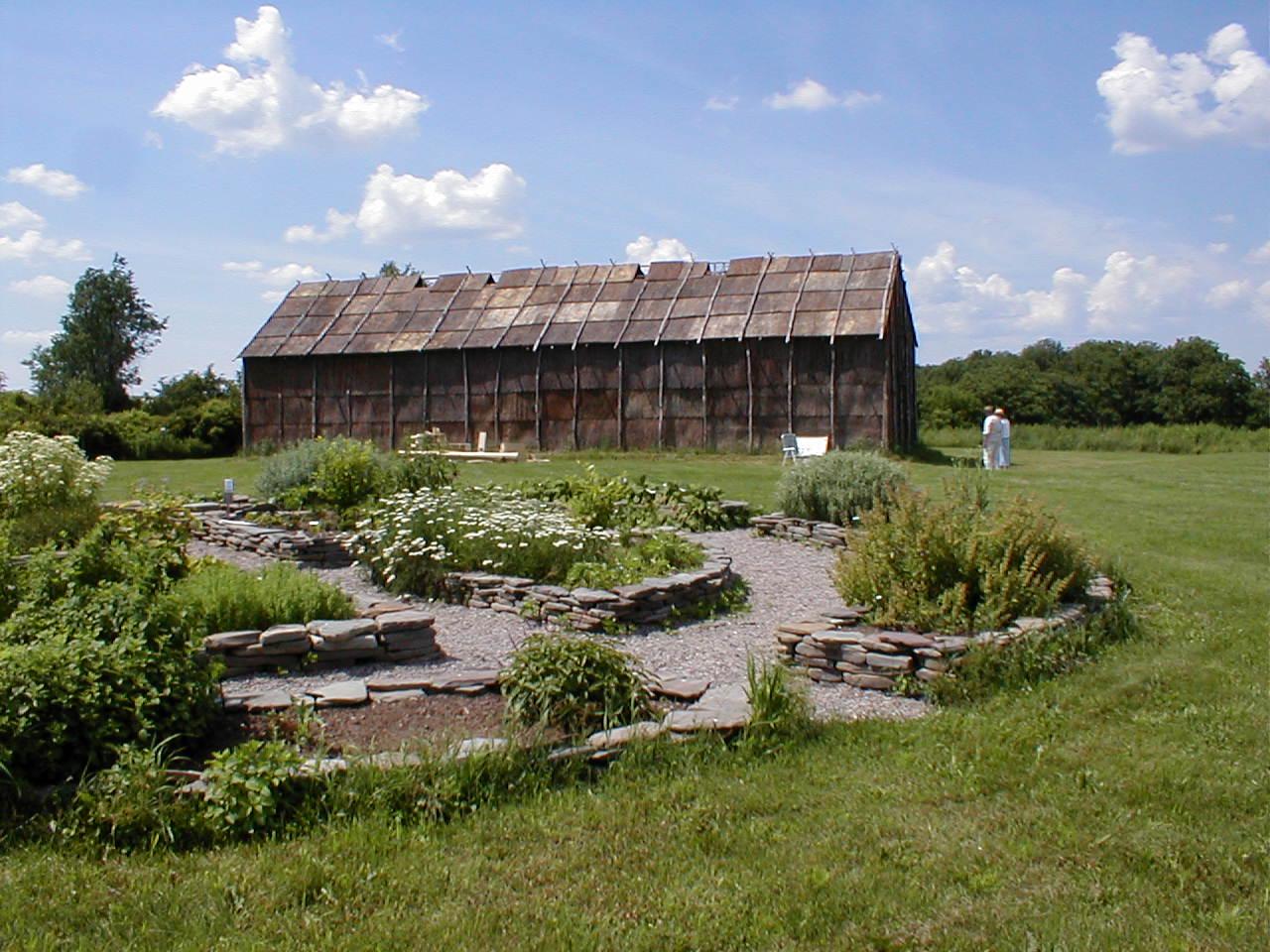 A long wooden building stands behind a stone-lined garden on a sunny day.