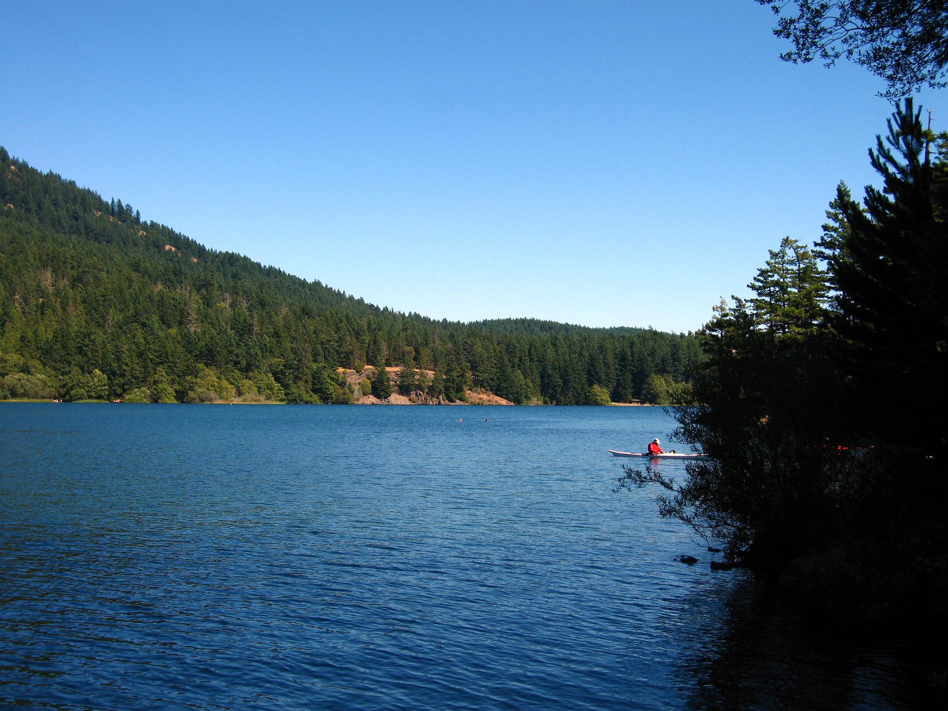 Sunny lake with forested hills, a kayaker near the shore.