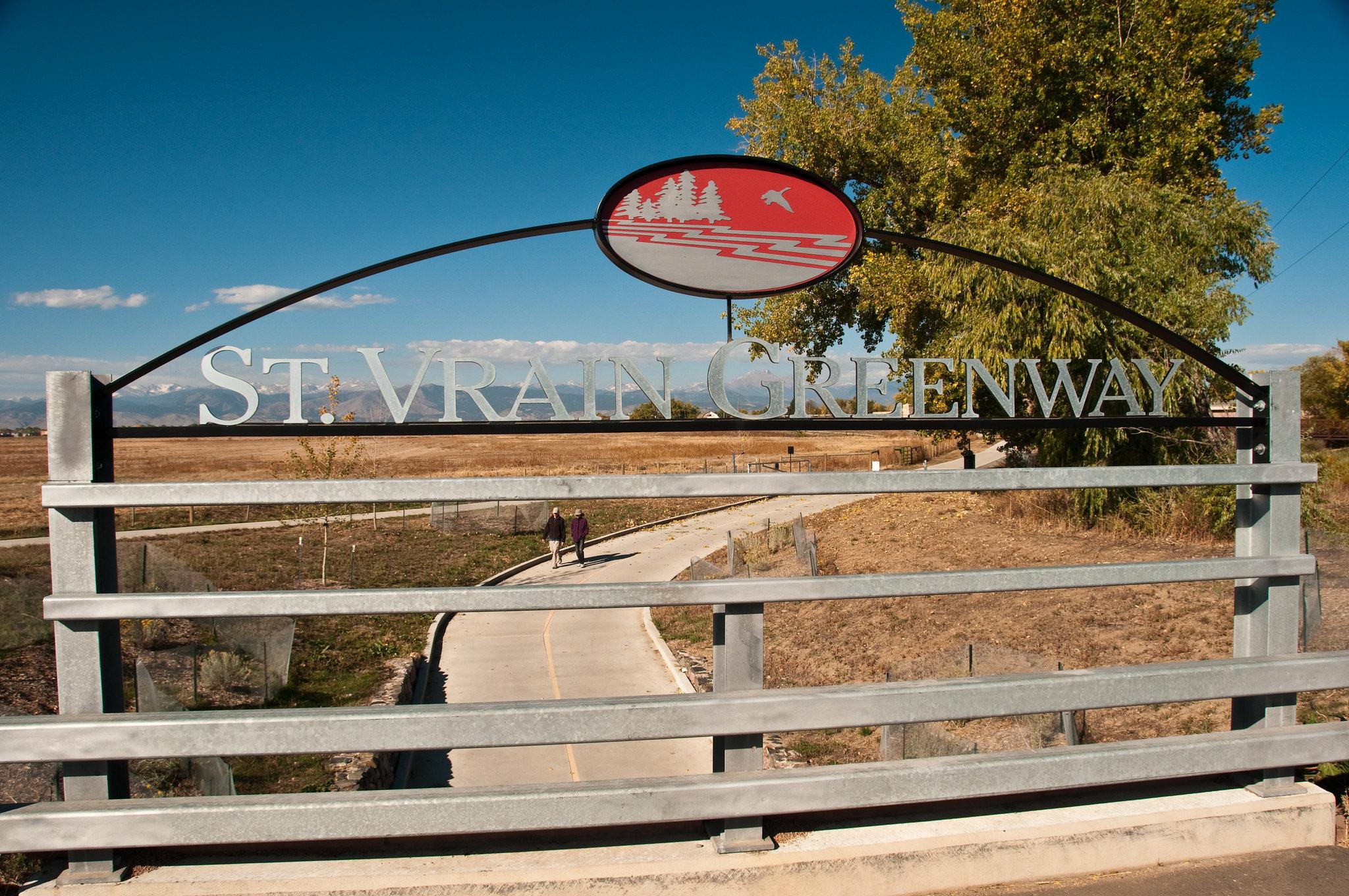 Metal archway sign over a sunny paved path with two distant walkers.