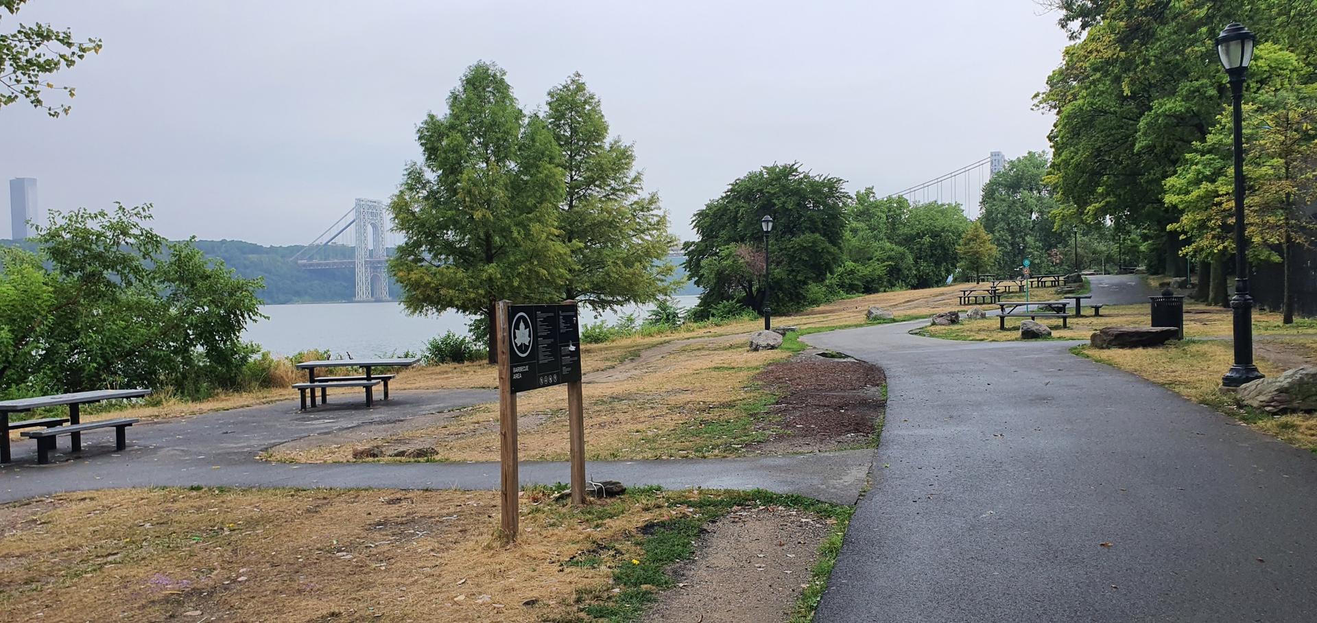 Park with picnic tables, paved path, and distant bridge under an overcast sky.
