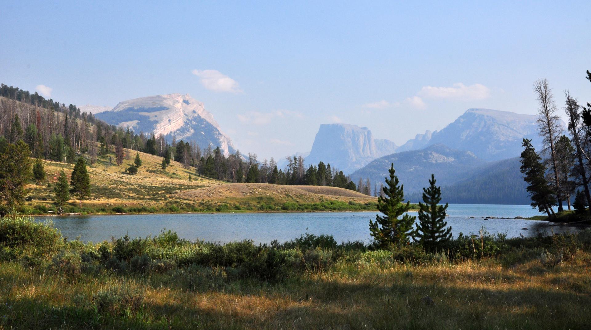 Calm lake reflecting bright sky, surrounded by forested hills and distant mountains.