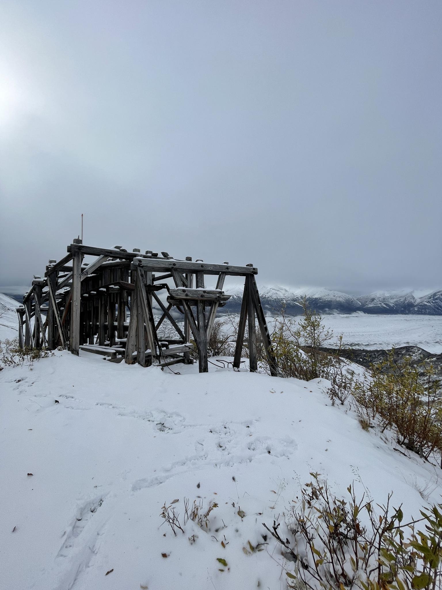 Old wooden structure on a snowy ridge overlooking a misty valley under overcast sky.