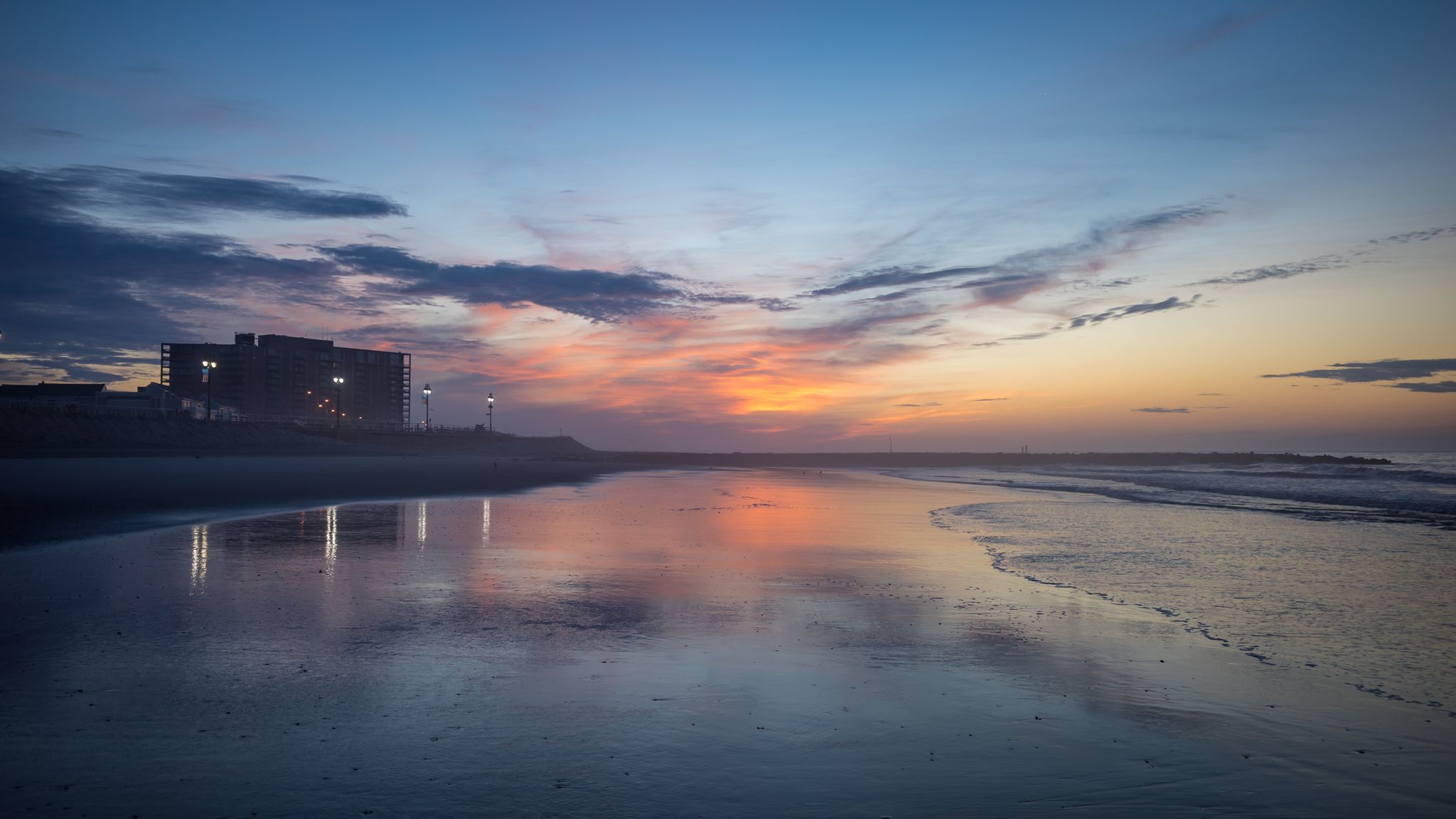 Vibrant sunrise over a calm beach, reflecting colorful sky and distant building. - Ocean City Boardwalk loop from Somers Point