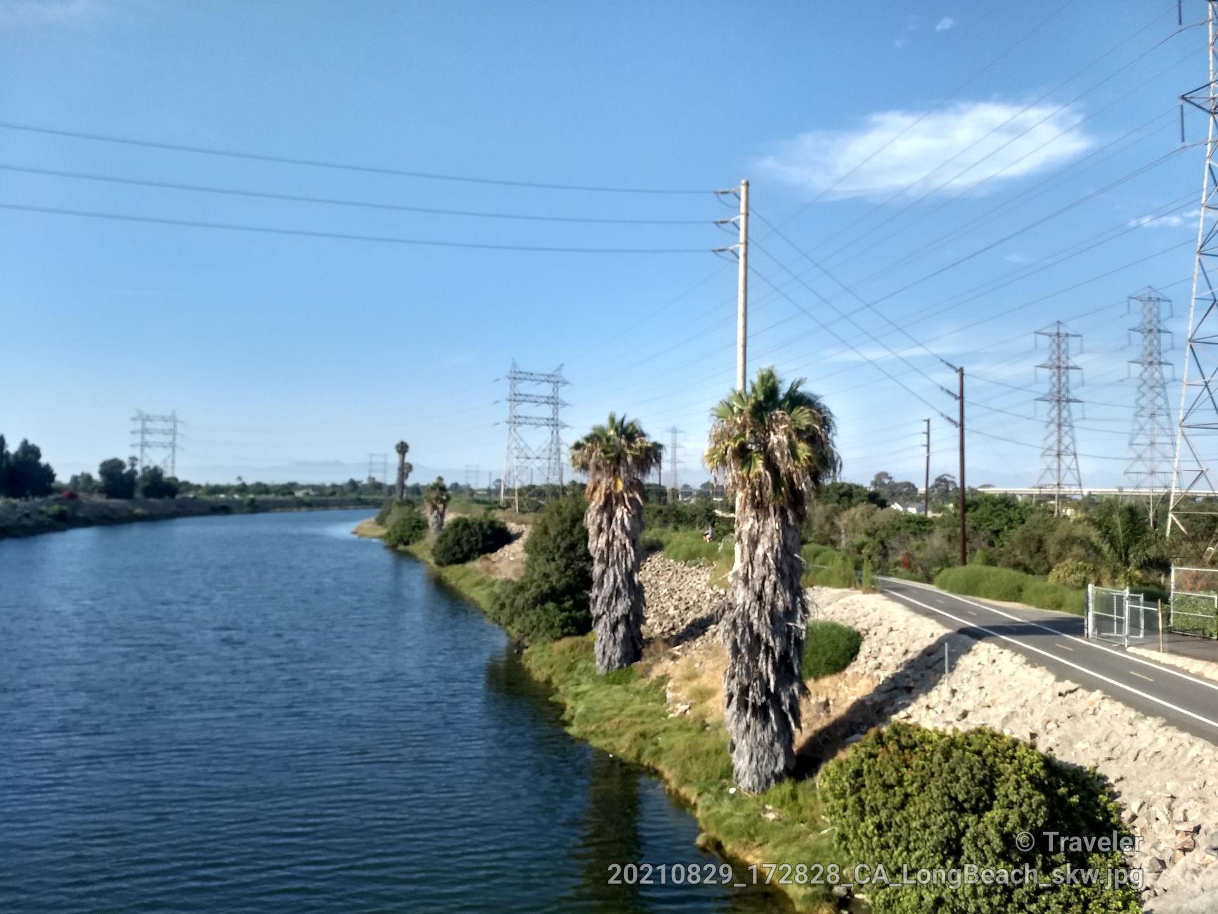 River flowing beside a paved path with palm trees and power lines.