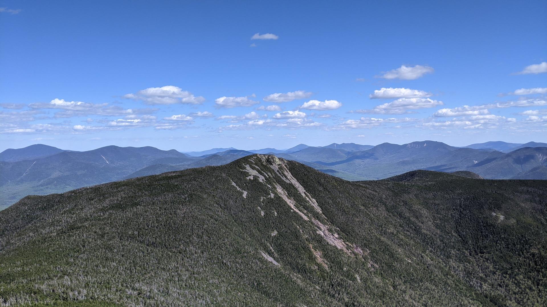 A forested mountain peak under a bright blue sky with scattered clouds.