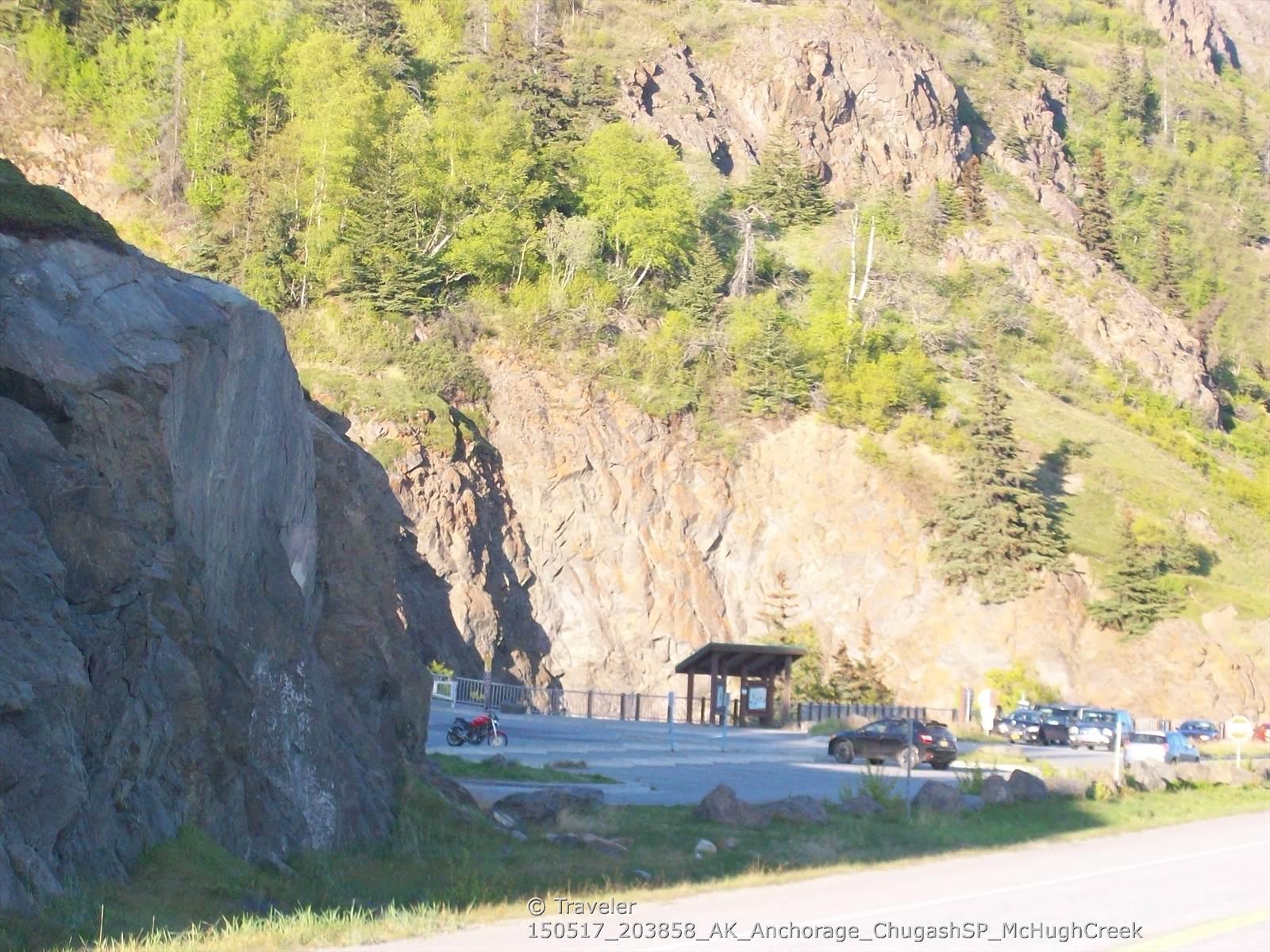 Cars parked in a lot at the base of a steep, rocky, forested mountain.