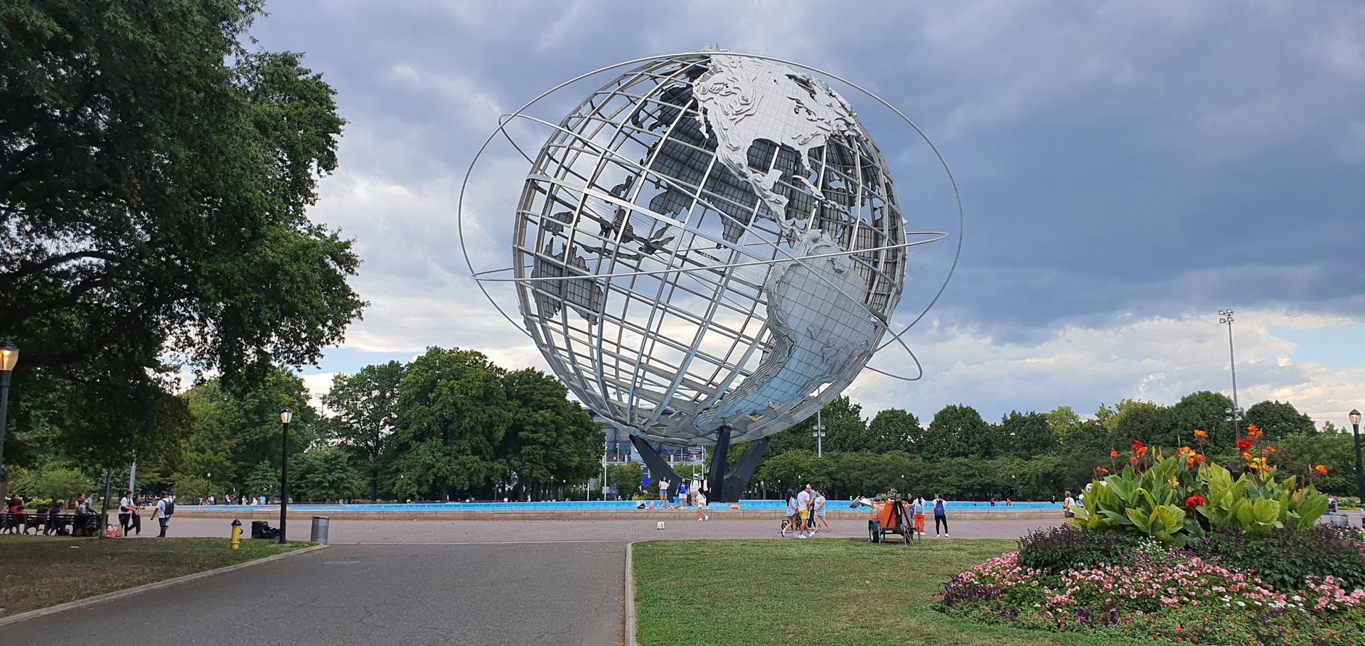 Large metallic globe sculpture in a park with people walking under cloudy skies.