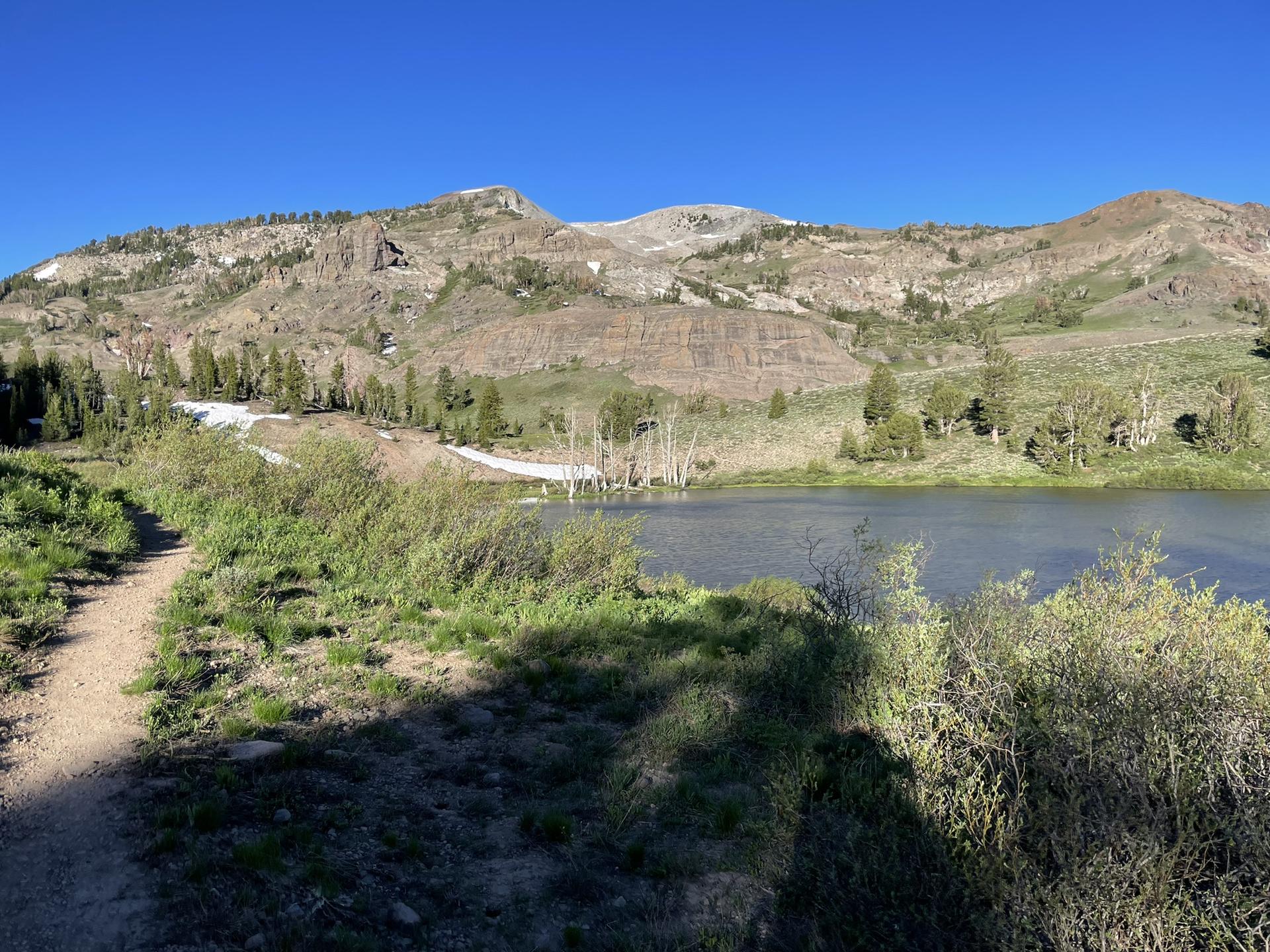A dirt path beside a lake with mountains and snow patches in bright sunlight.