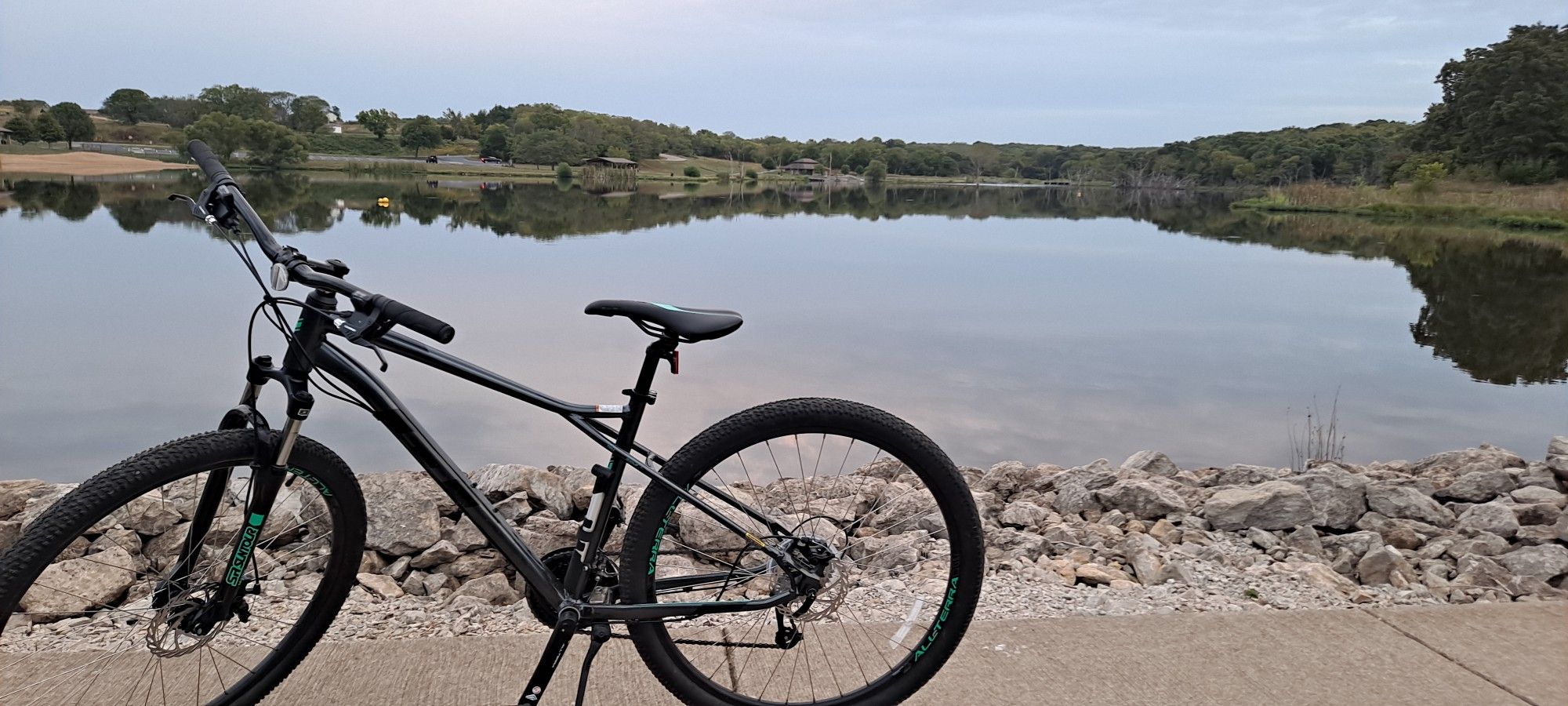 Black bicycle parked on a paved path beside a calm lake under an overcast sky. - Kill Creek Streamway & Cedar Niles Trail loop