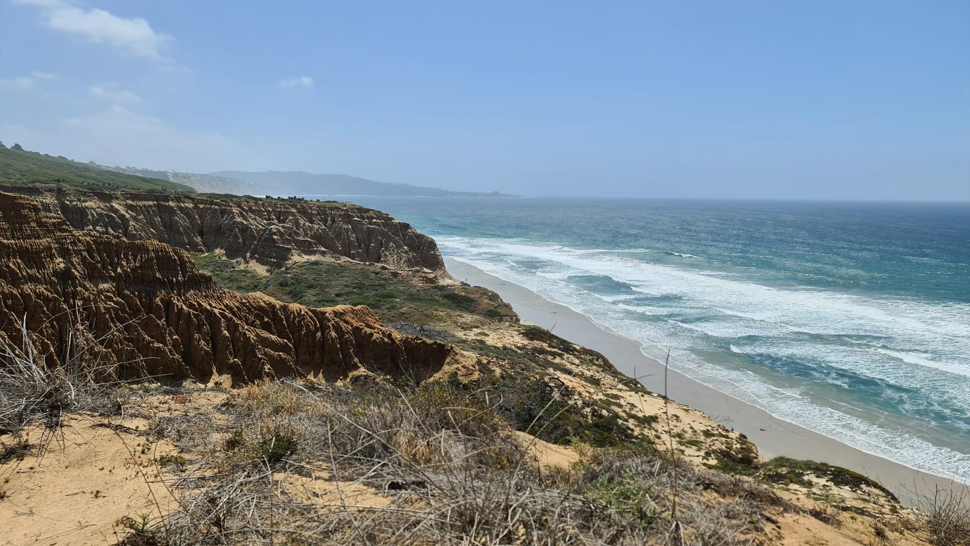 Sunny expansive view of sandy beach, ocean waves, and coastal cliffs.