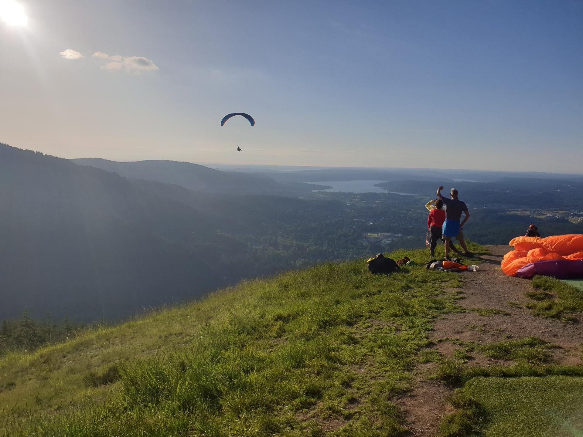 A paraglider soars over a sunny, grassy hill with people watching.