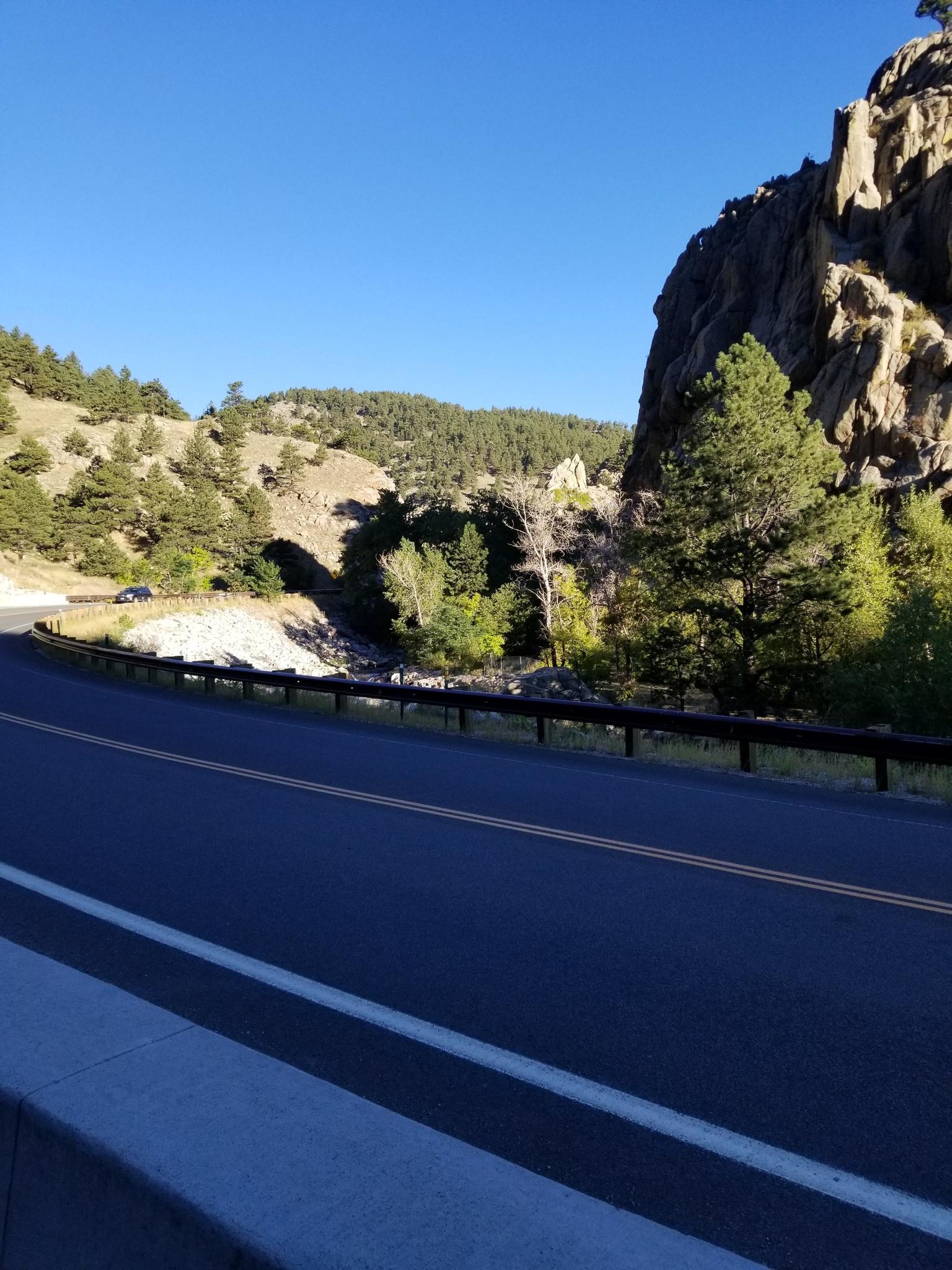 A winding paved road curves beside a rocky cliff and forested river valley.