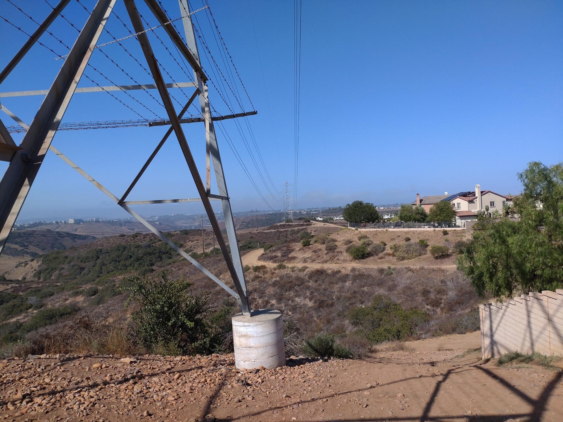 A large power line tower stands on a sunny, dry hillside with distant houses.