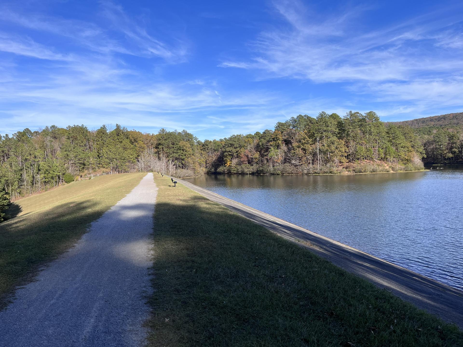 Wide gravel path beside a calm lake with forested hills under blue sky.