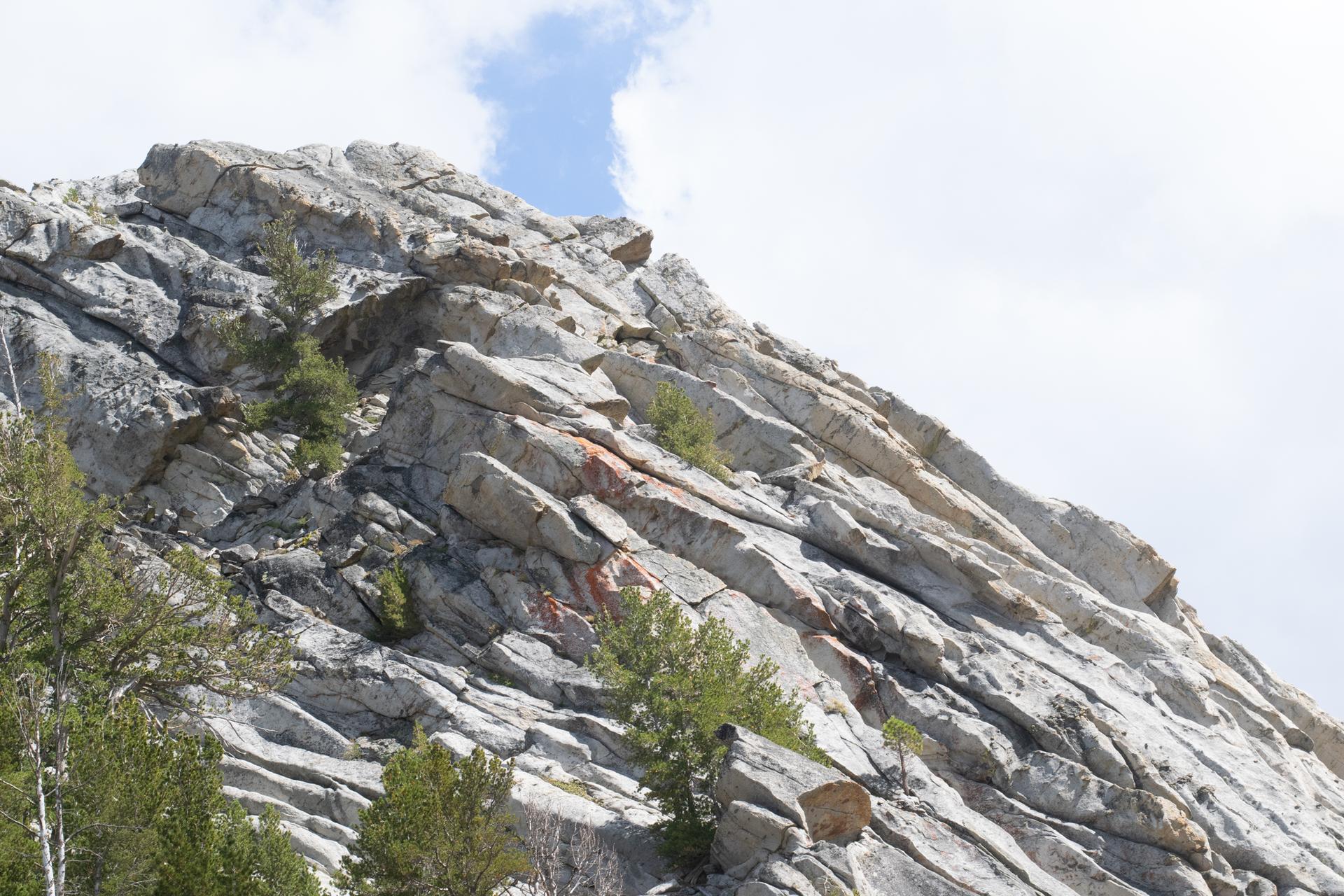 A steep, rugged rock face with sparse green trees under a cloudy sky.