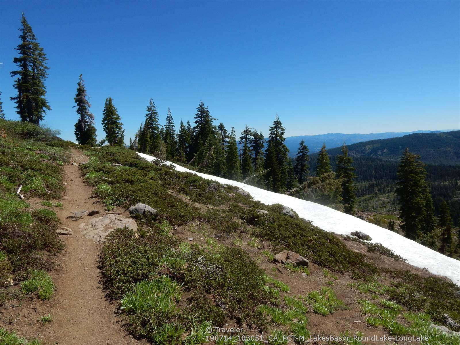 Dirt trail next to snow on a hillside with pine trees and distant views.