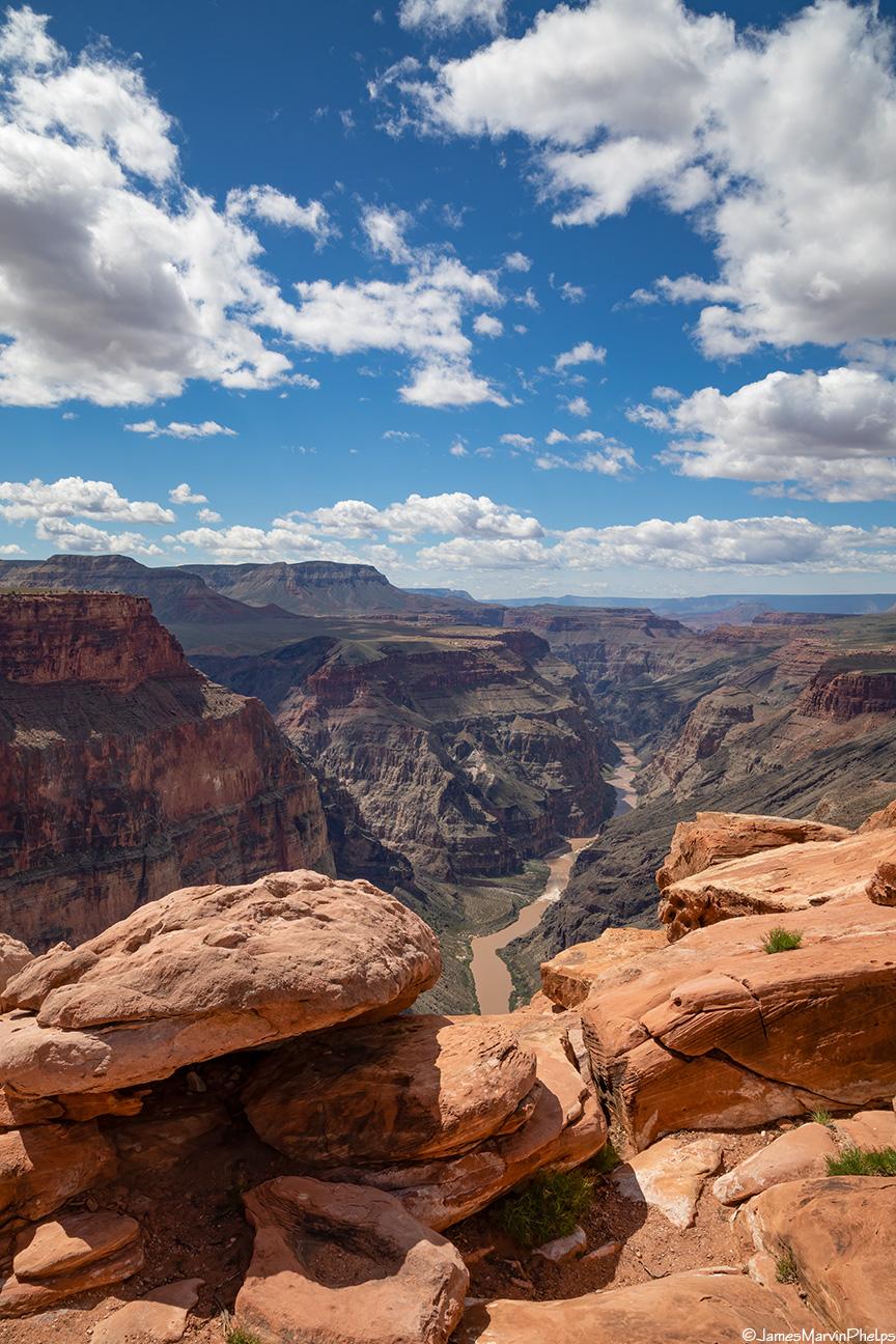 Deep canyon vista with a river, layered rock, and bright, cloudy sky.