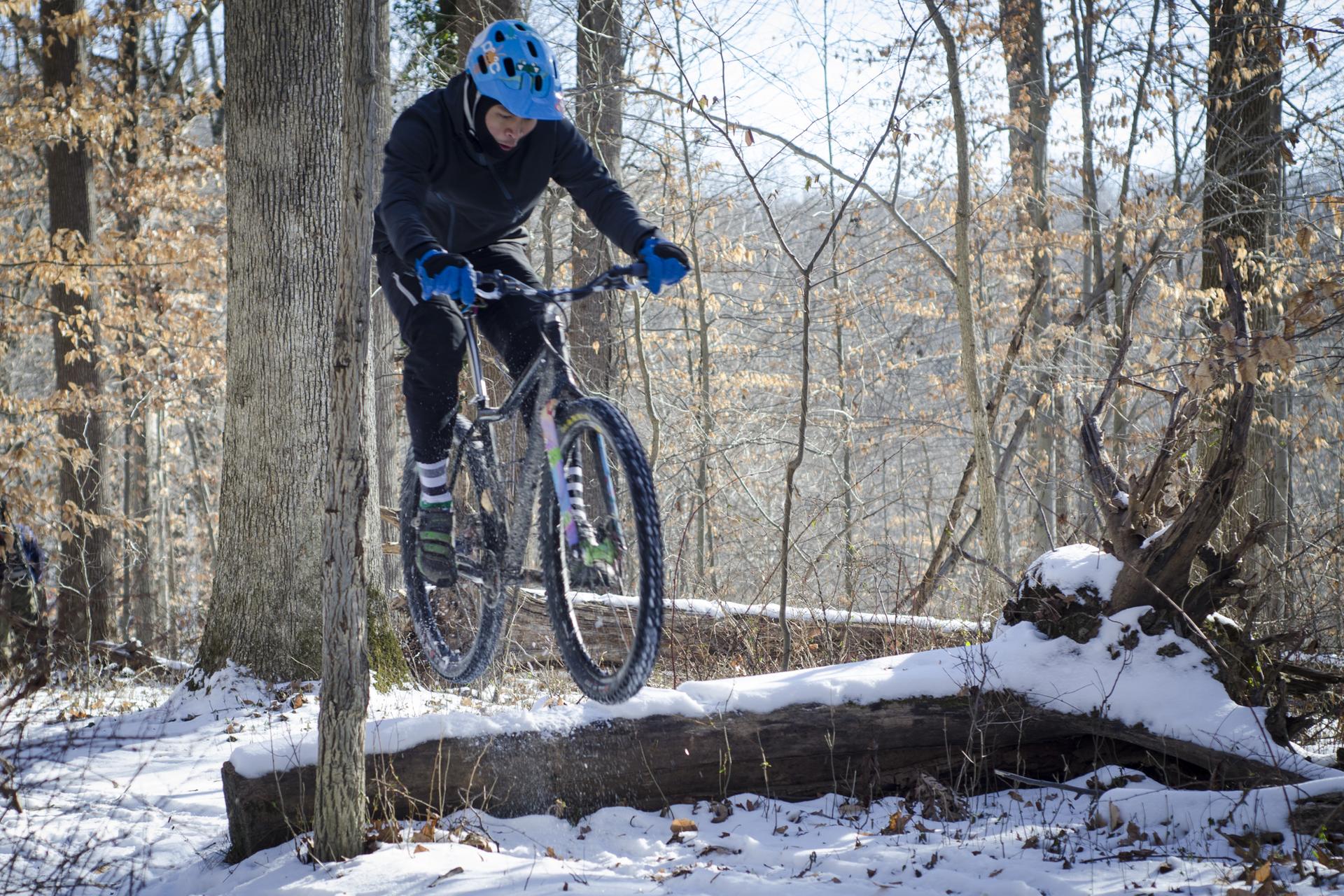 Person on a mountain bike jumping over a snowy log in a forest.