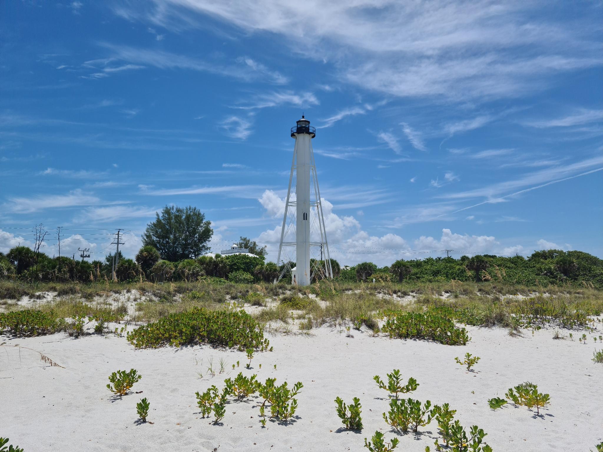Tall white lighthouse on a sandy beach under a bright blue sky.