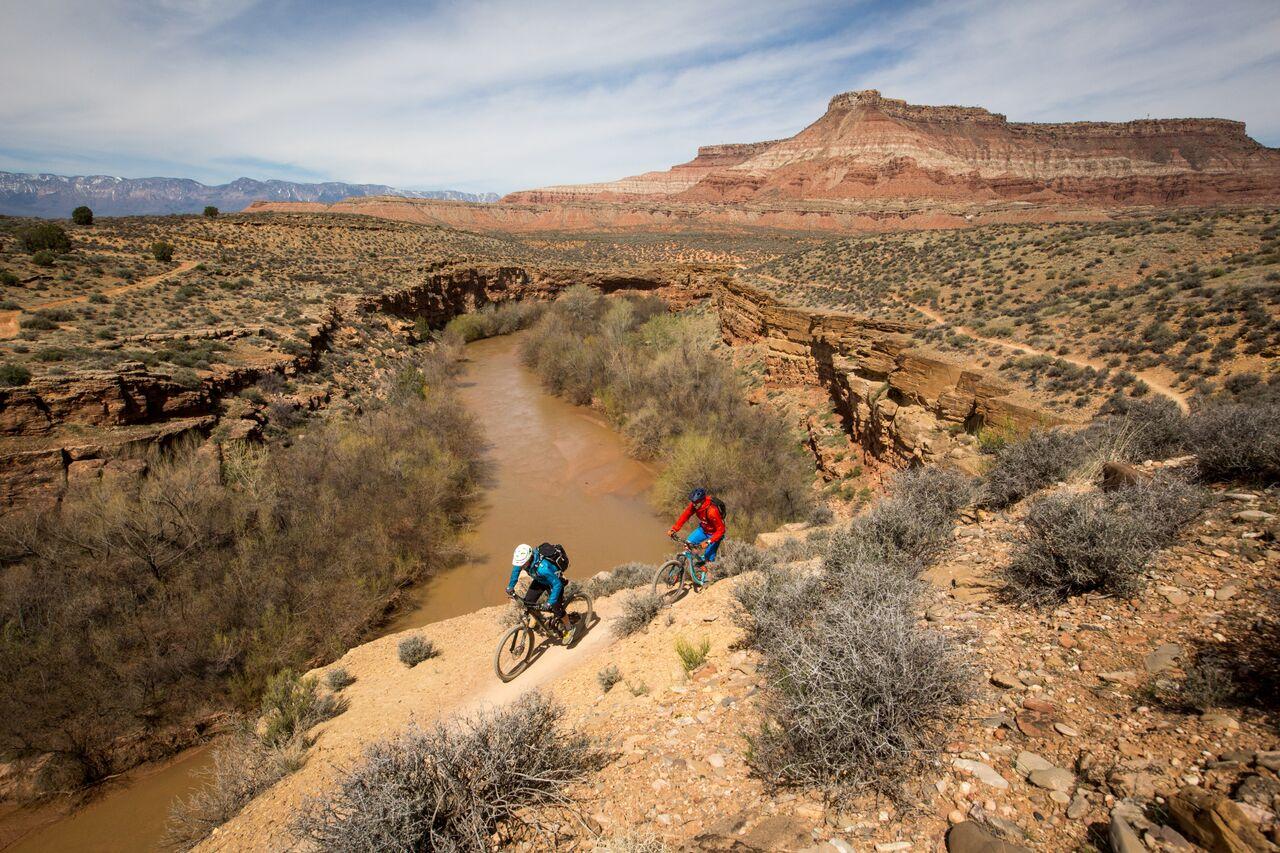 Two mountain bikers ride a desert trail beside a muddy river under bright skies.