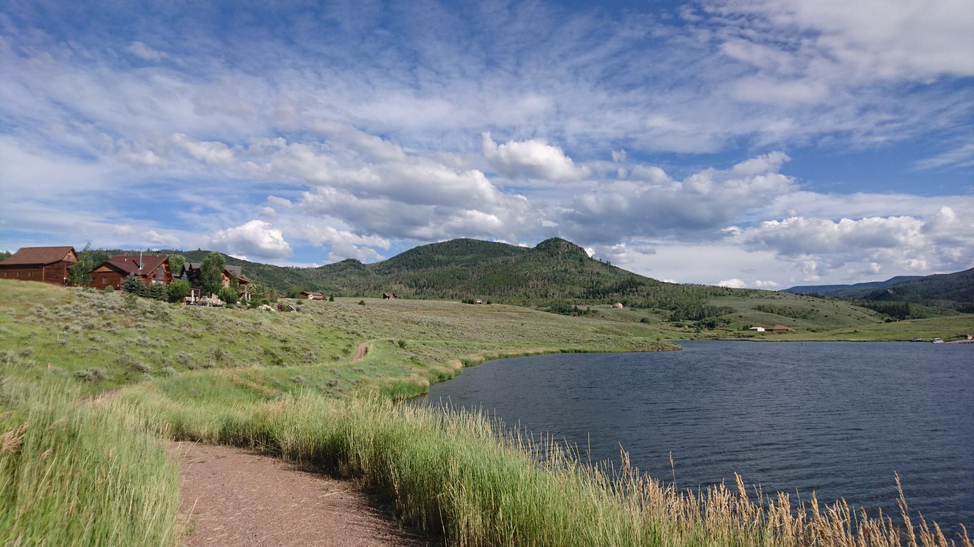 A grassy path next to a lake, with distant houses and mountains under a cloudy sky.