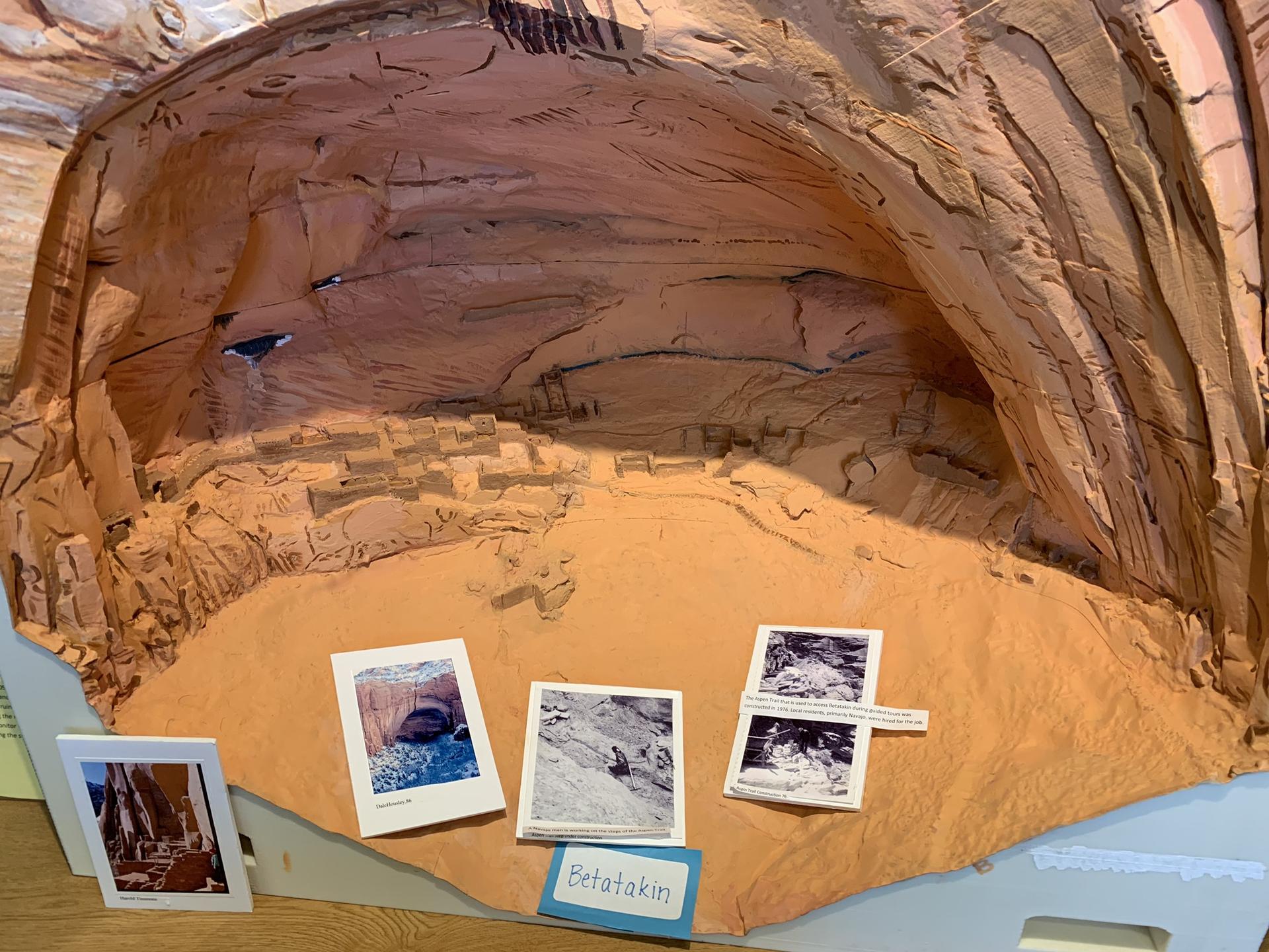 Diorama of an ancient cliff dwelling with small photos on the sandy ground.