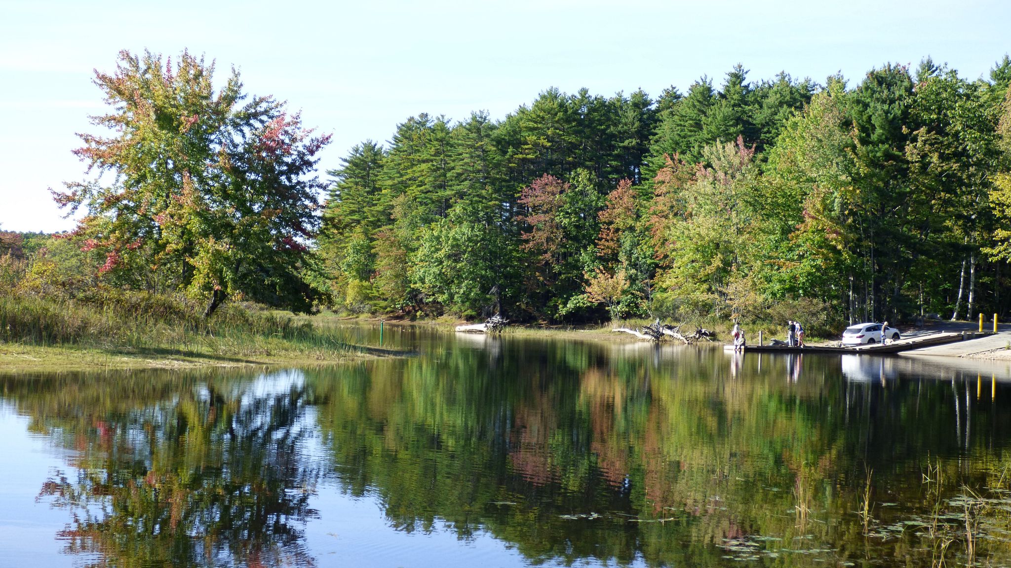 Calm water reflects autumn trees and a boat launch with people in bright daylight. - Outer Loop
