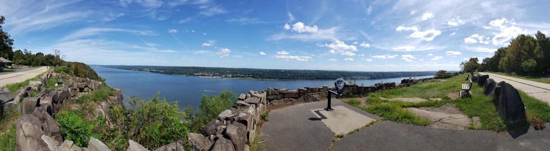 Panoramic view of a wide river and distant land under a bright, cloudy sky.