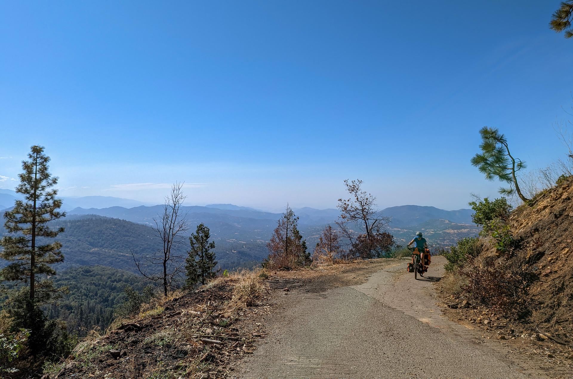 Cyclist on a mountain road overlooking a vast, hazy valley under clear sky.